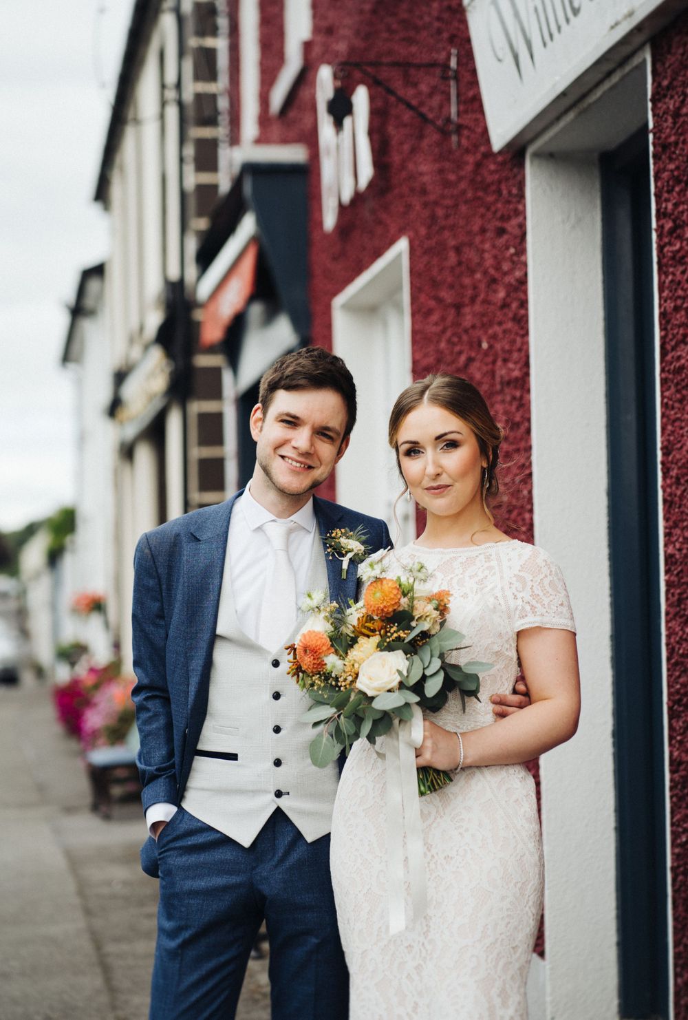 A couple poses outside red and white buildings, with the bride holding an orange and white bouquet.