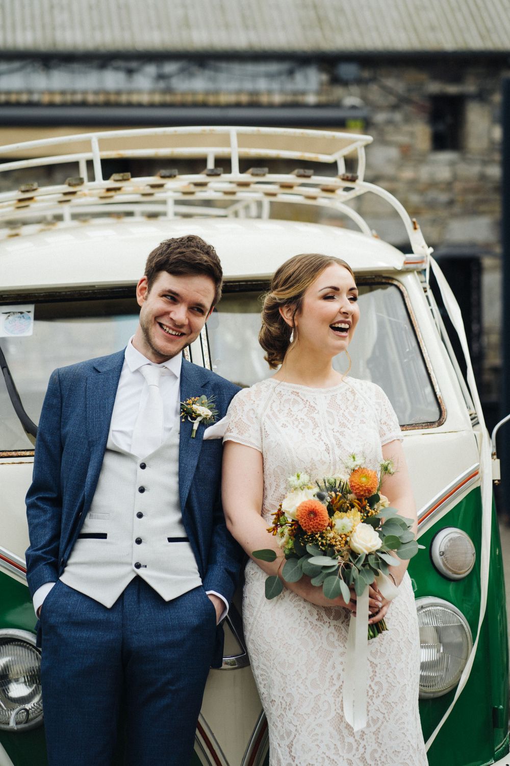 A happy couple laughs while posing next to a classic green and white VW van on their wedding day.