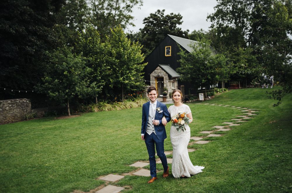 A couple walking along a stone path through a lush green garden at an outdoor wedding venue.