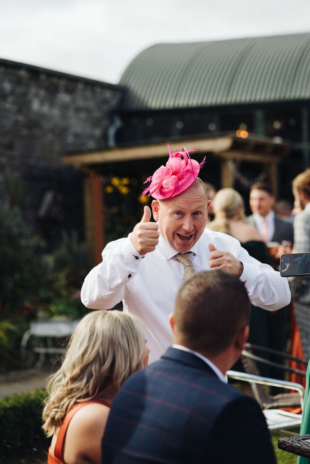 Wedding guest in white shirt and pink fascinator gesturing enthusiastically at an outdoor reception.