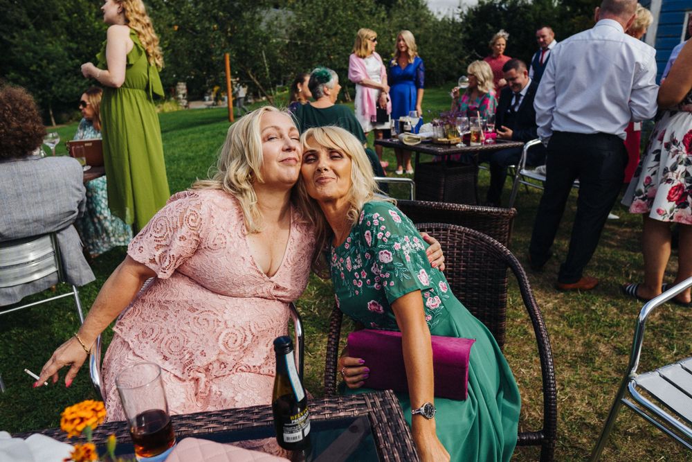 Two wedding guests sharing a joyful moment on outdoor furniture at a garden reception.