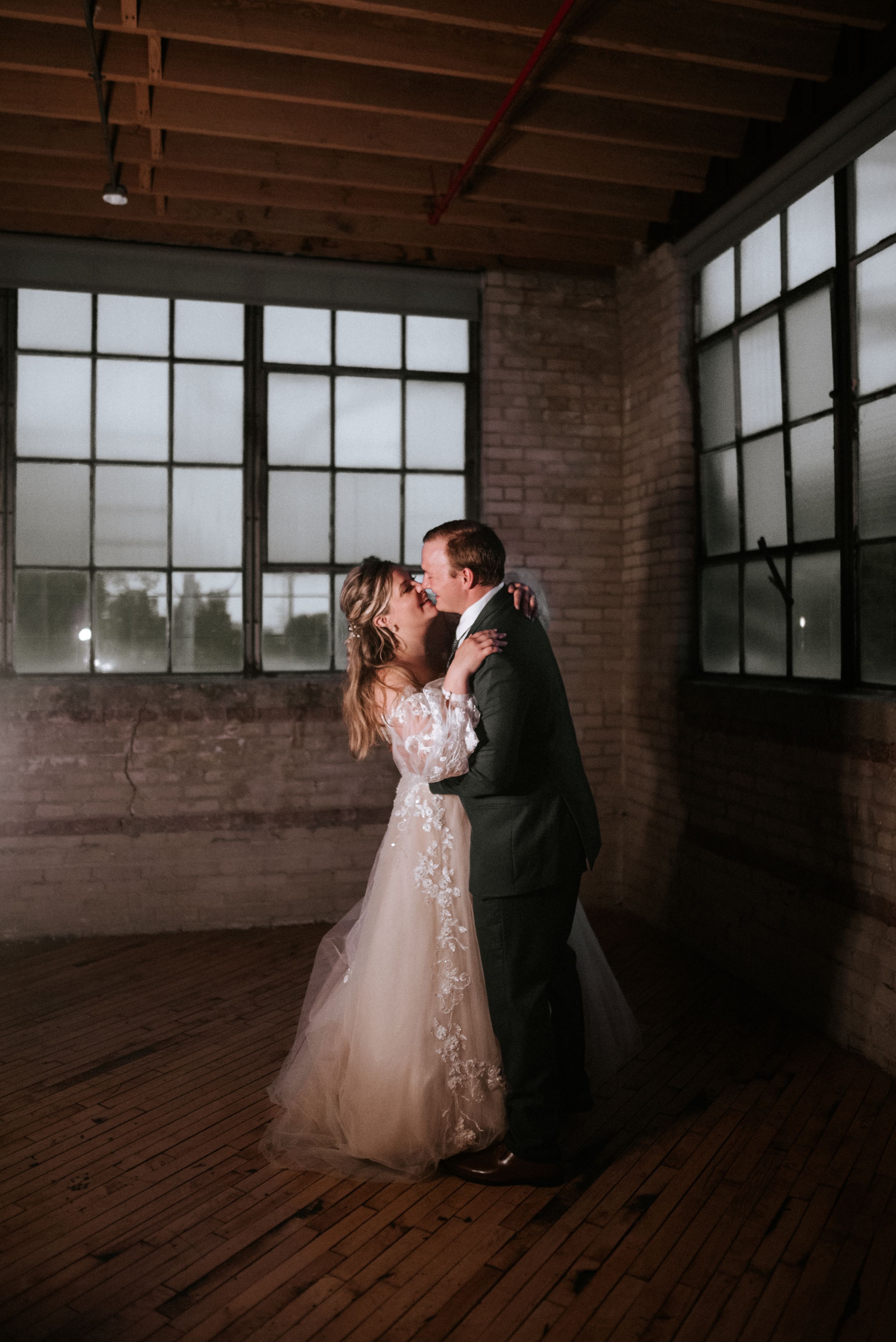 A romantic dance moment in a rustic brick venue with large windows and wooden floors.