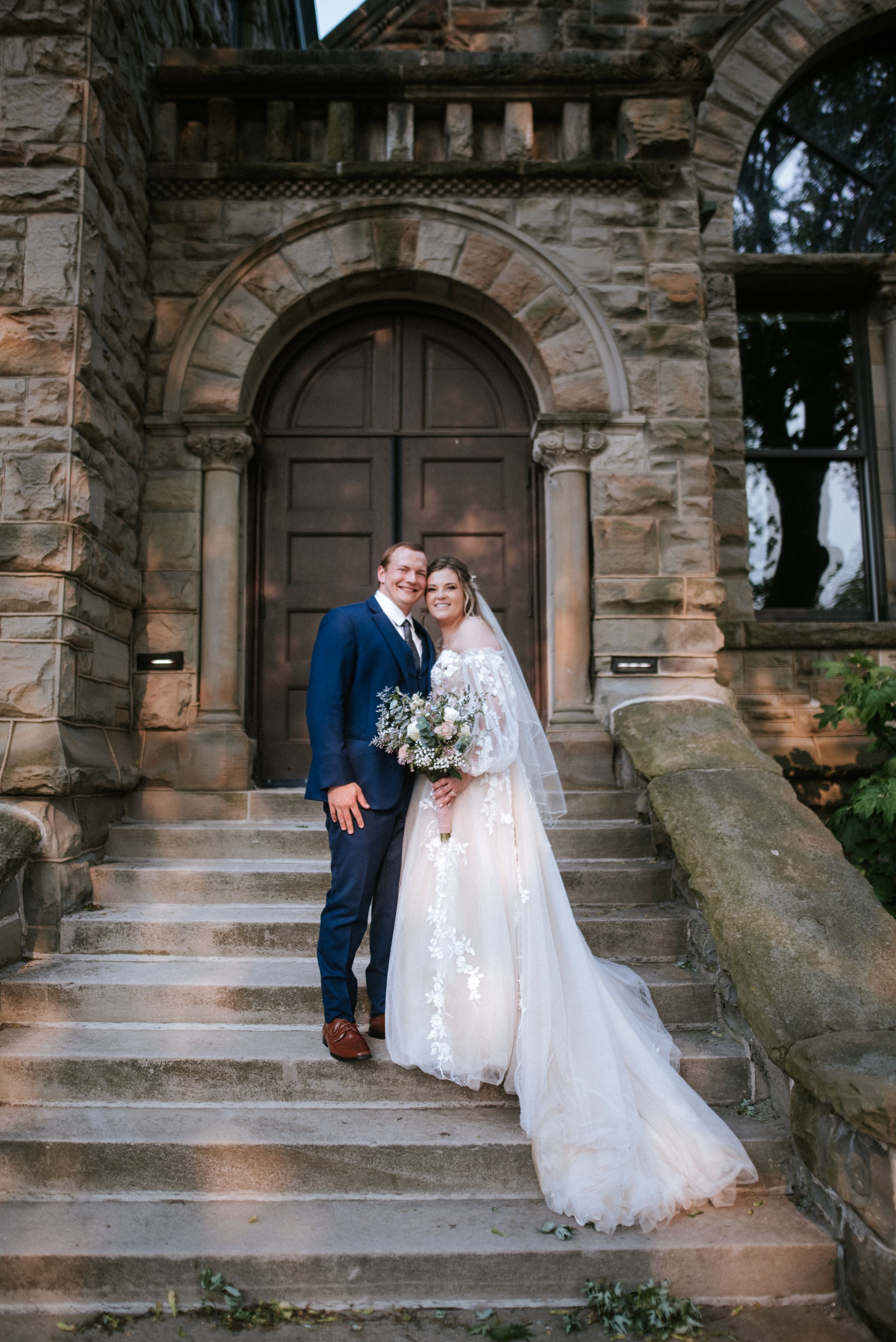 Couple poses on stone steps of historic arched doorway with trailing wedding dress.