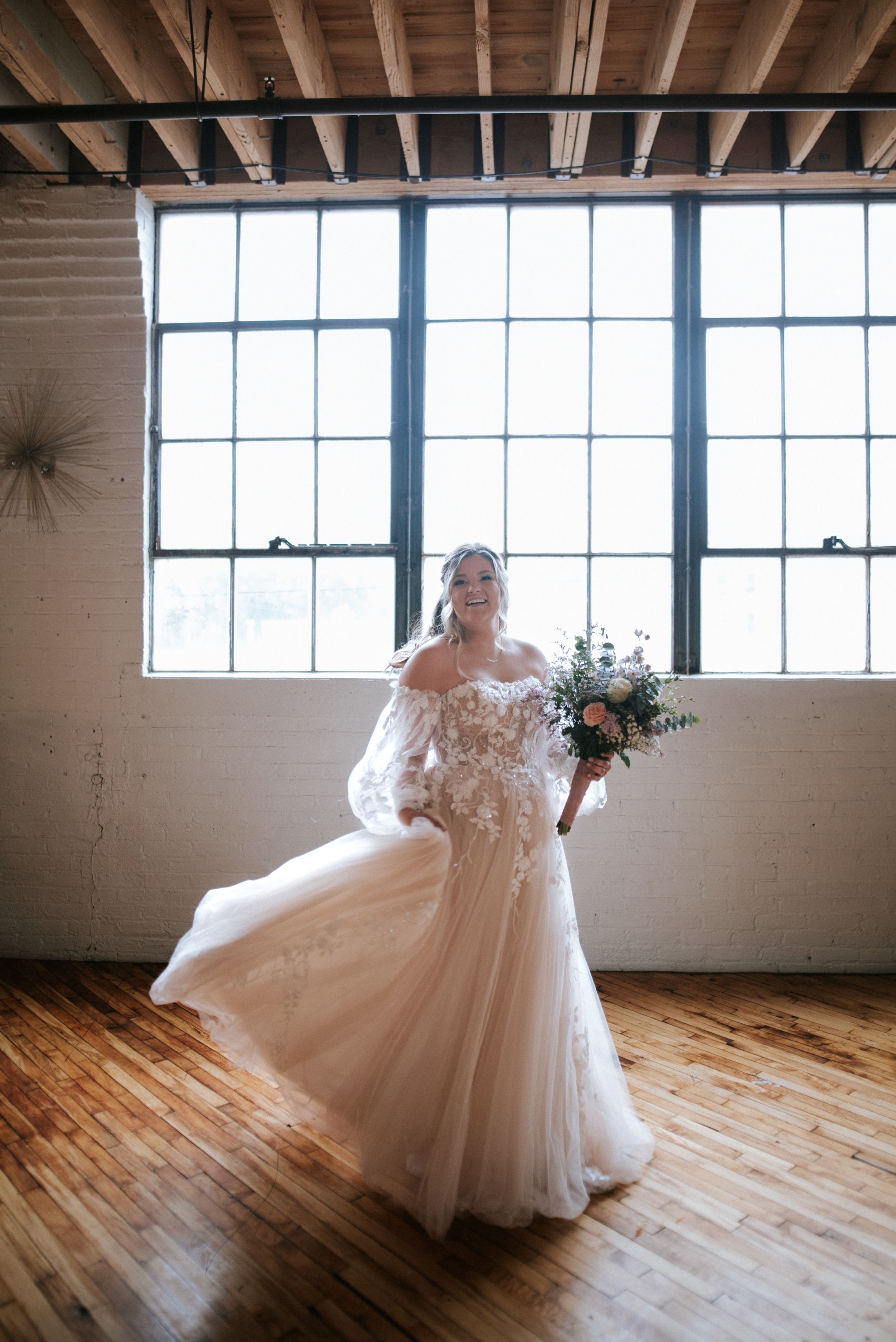 Bride in off-shoulder lace gown twirls in rustic loft space with large windows.