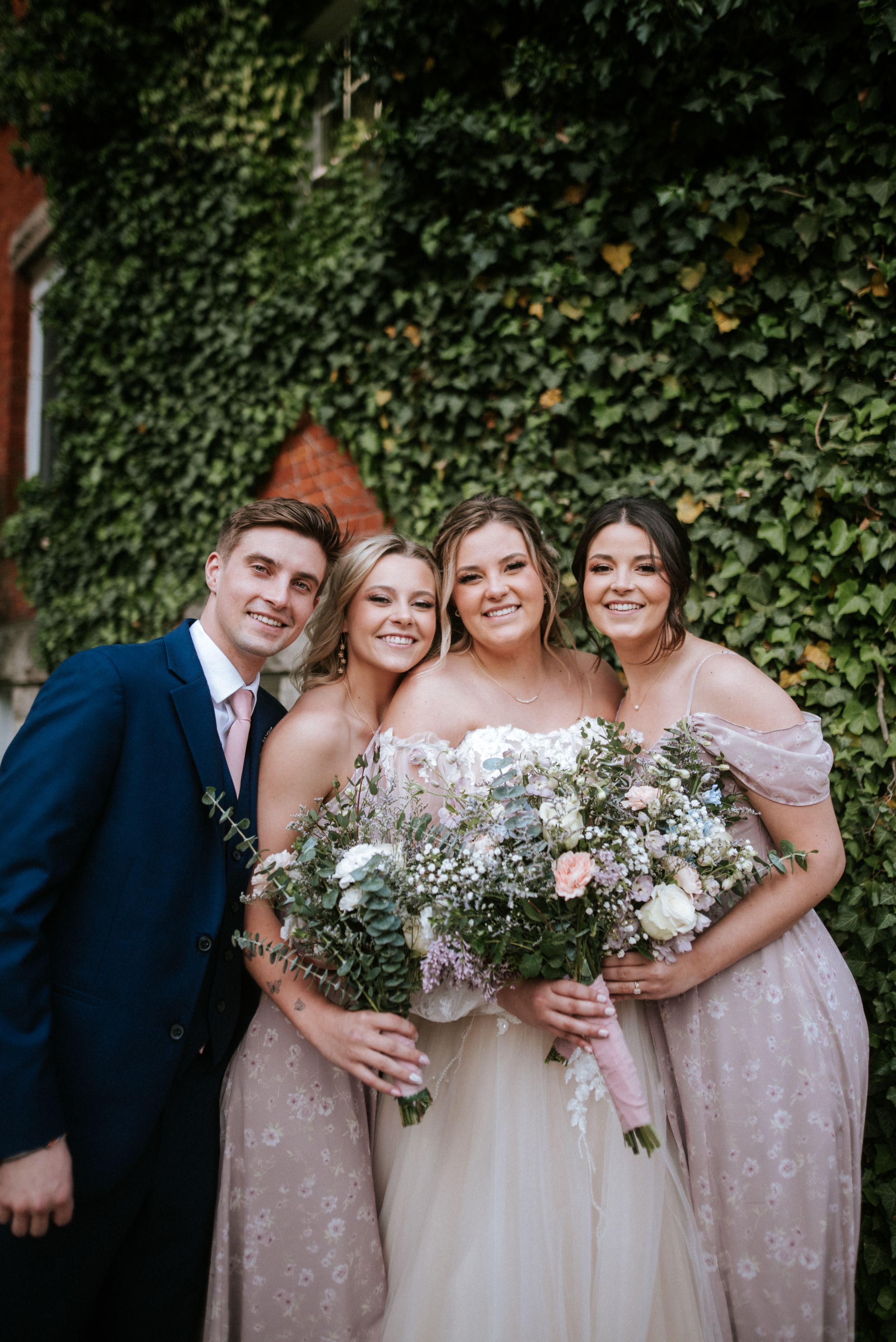 Small wedding party group poses with floral bouquets against ivy-covered wall.