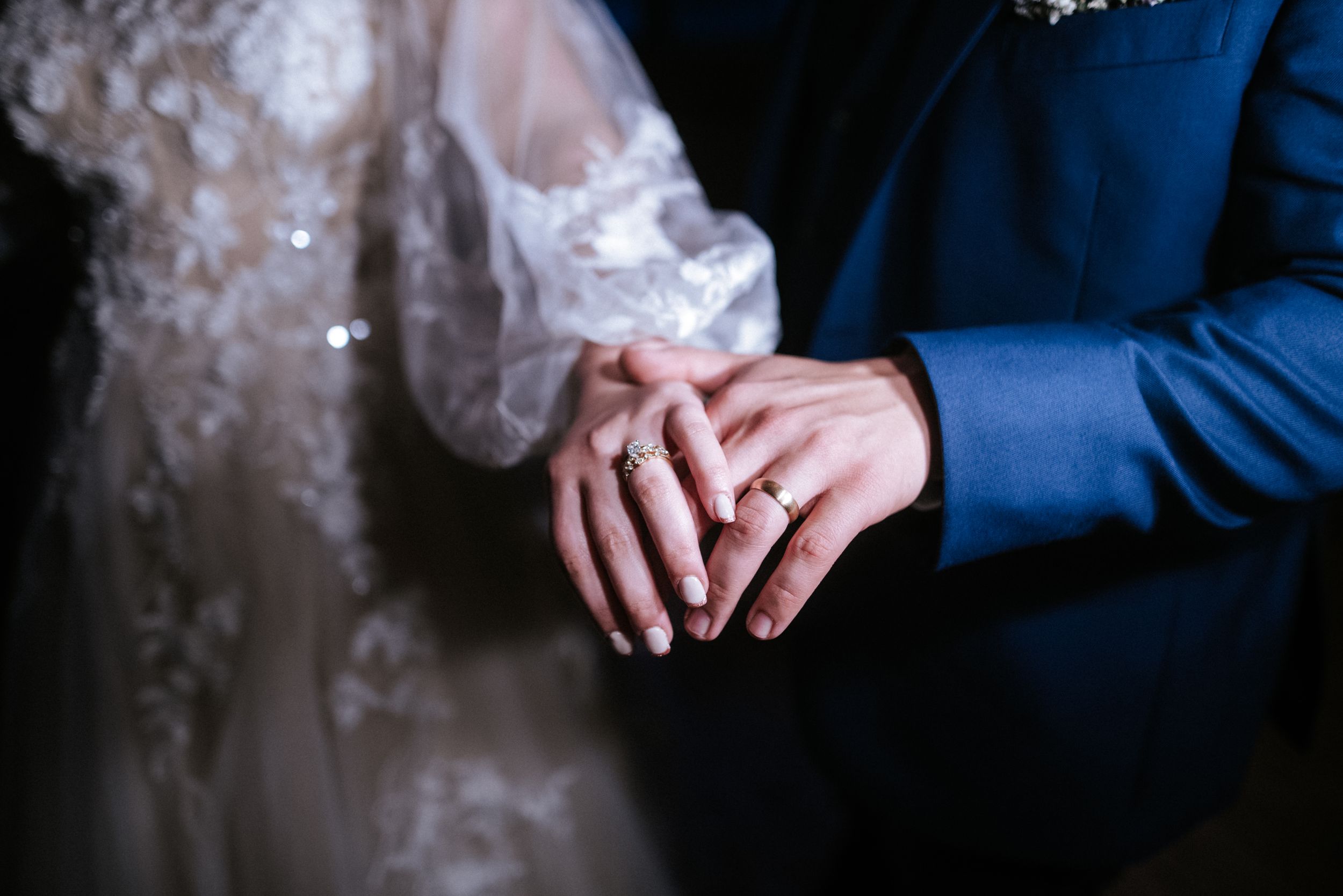 Close-up detail shot of wedding rings and lace dress against a navy blue suit.
