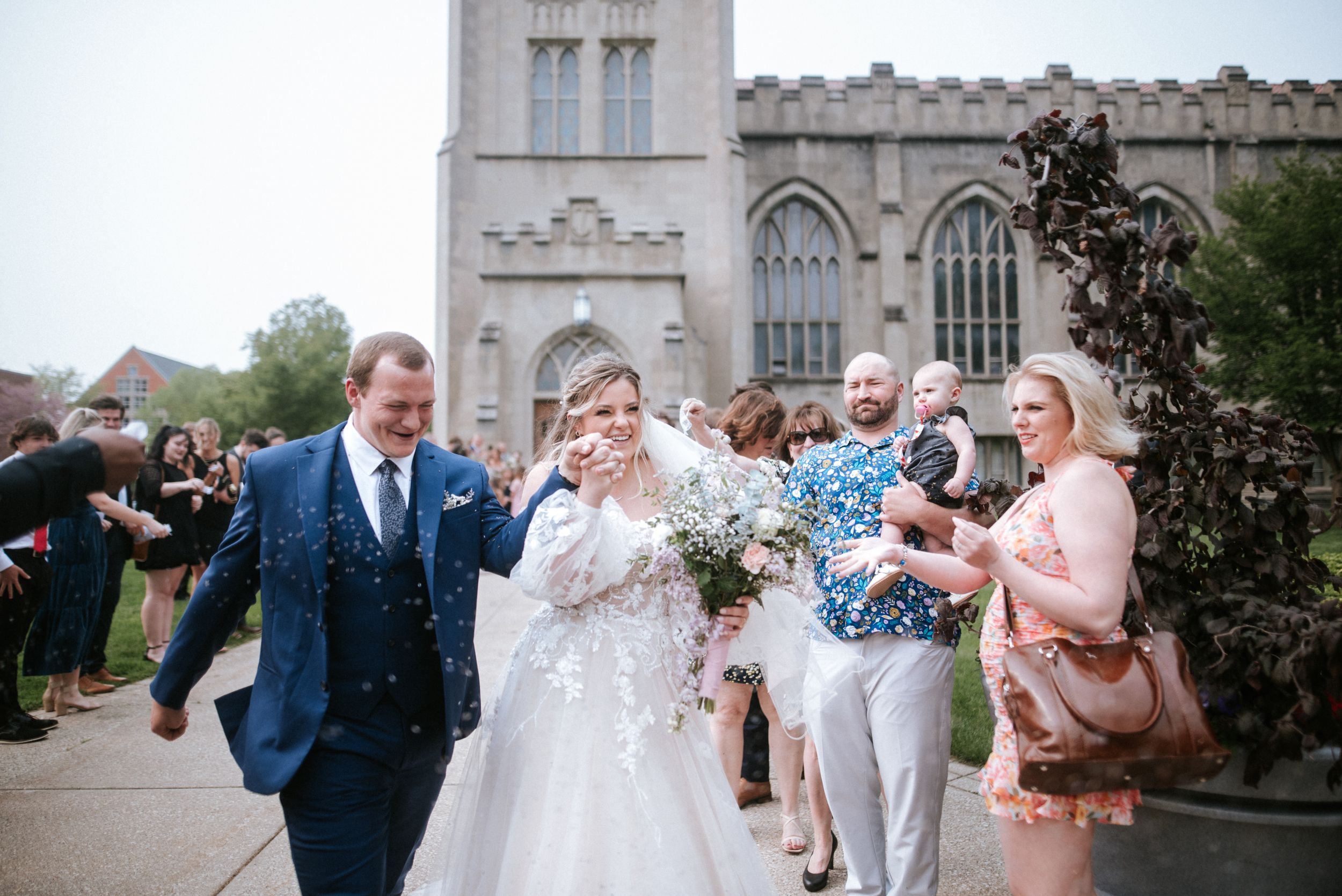 Joyful couple walks past celebrating guests outside stone church during wedding exit.