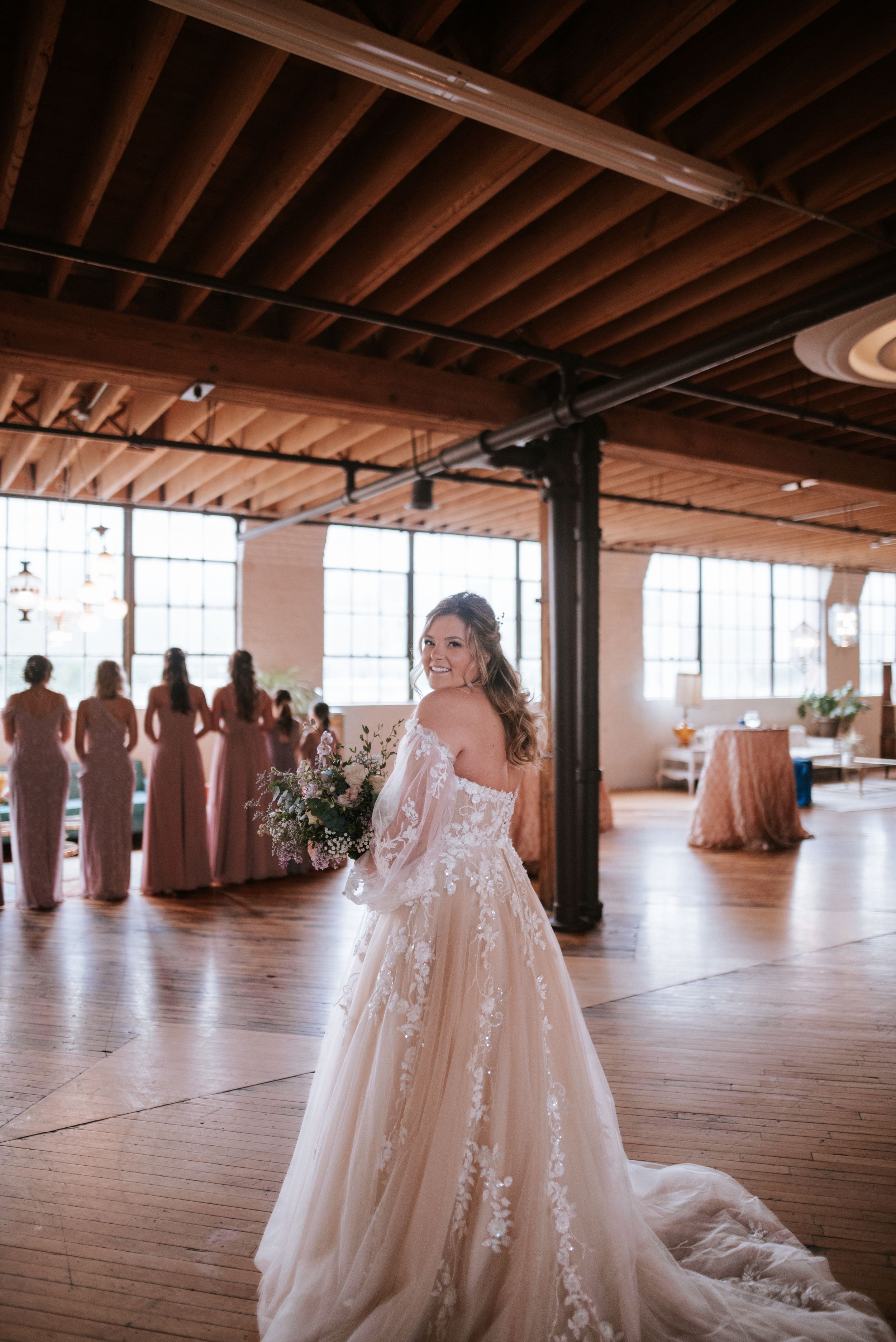 Bride in lace gown stands in rustic venue with bridesmaids in background.