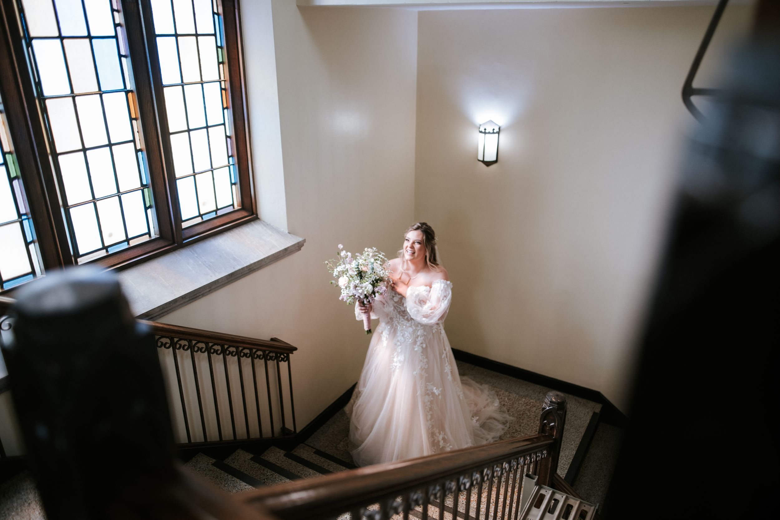 A bride in a white lace gown holds a bouquet while standing on an elegant staircase near a leaded glass window.