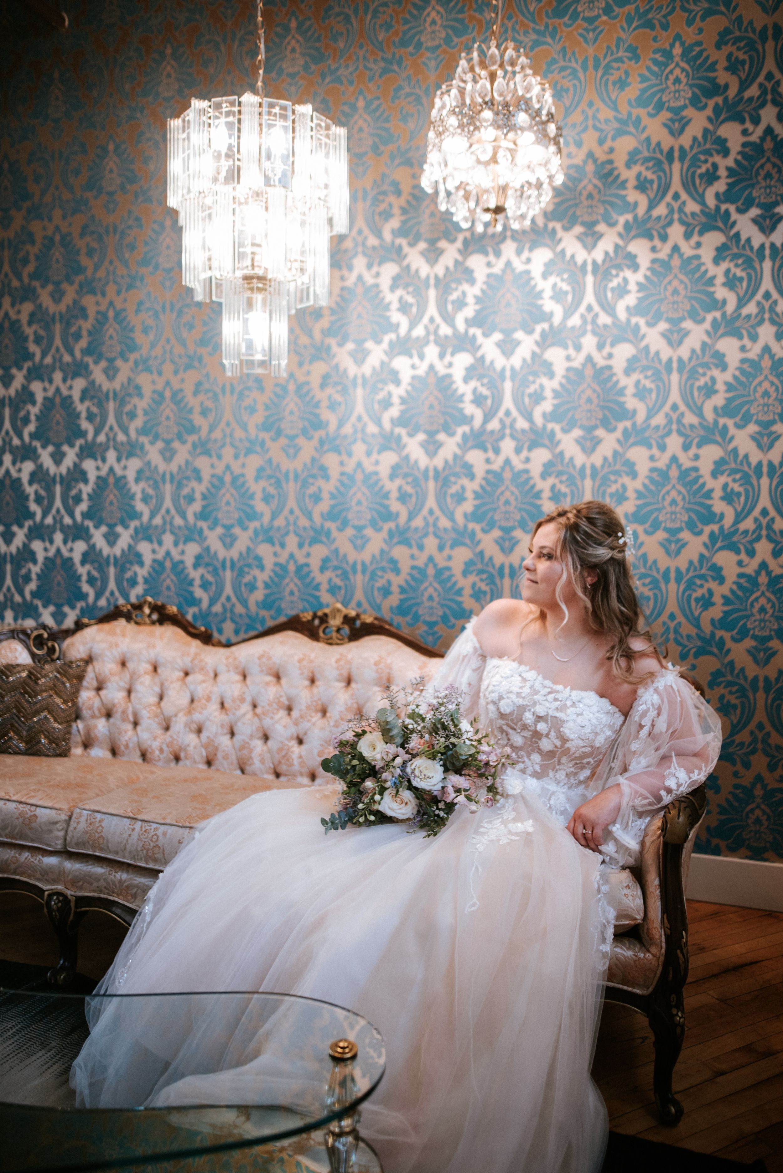 Bride in white gown sits on vintage sofa under crystal chandeliers with elegant wallpaper background.