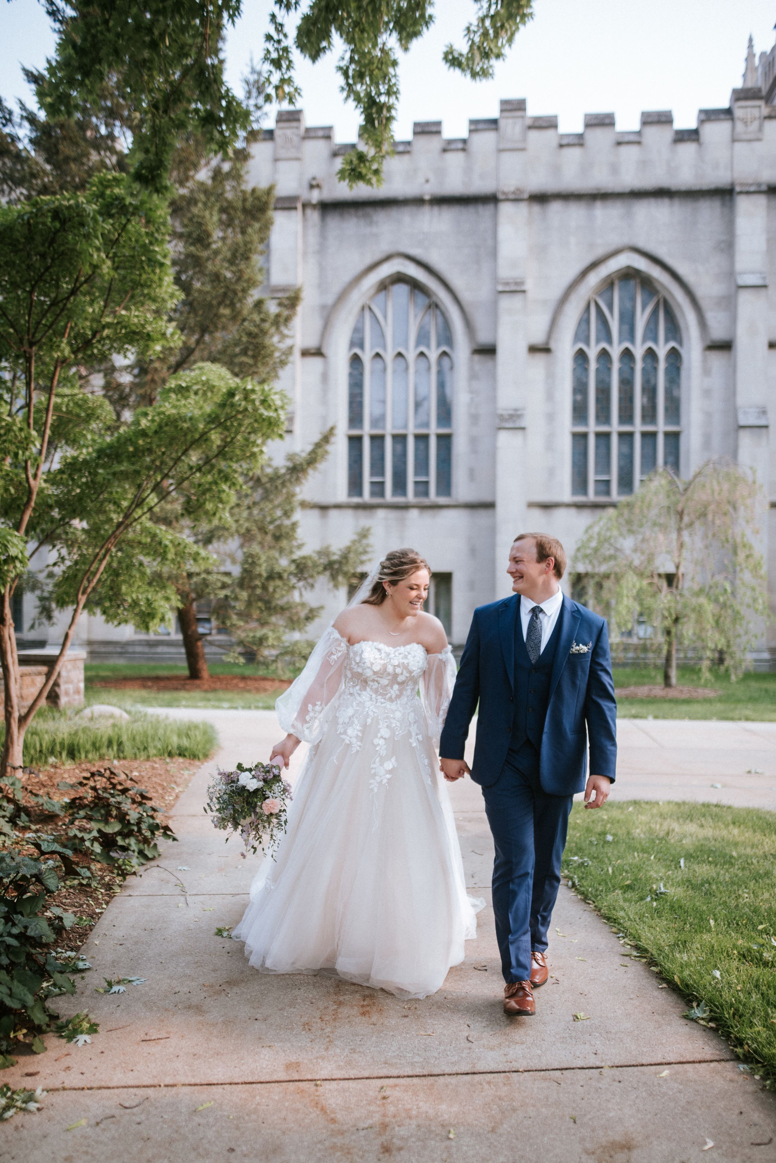 Couple walks hand in hand along garden path beside gothic church windows.