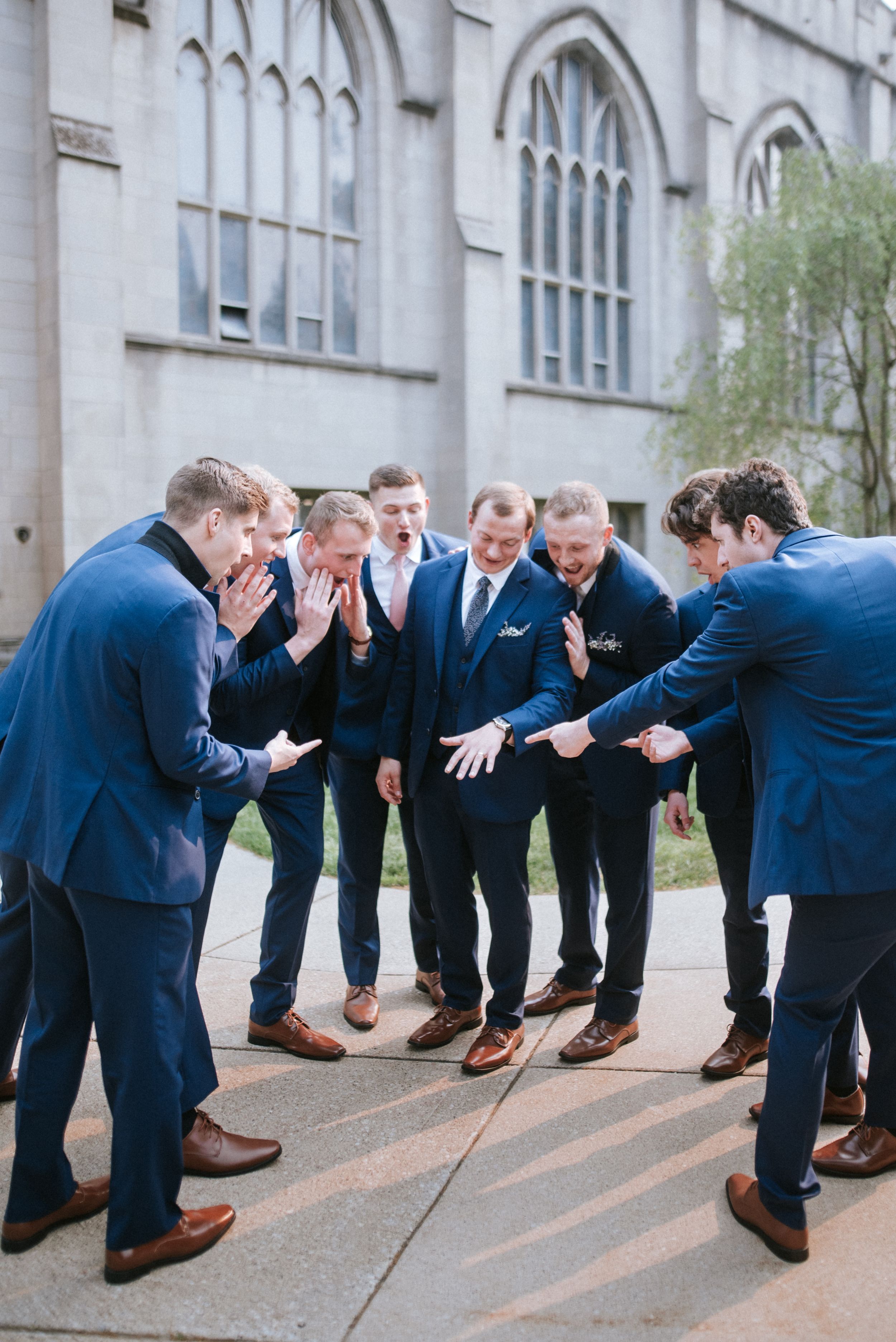 Groomsmen in navy suits huddle in playful pose outside gothic church building.