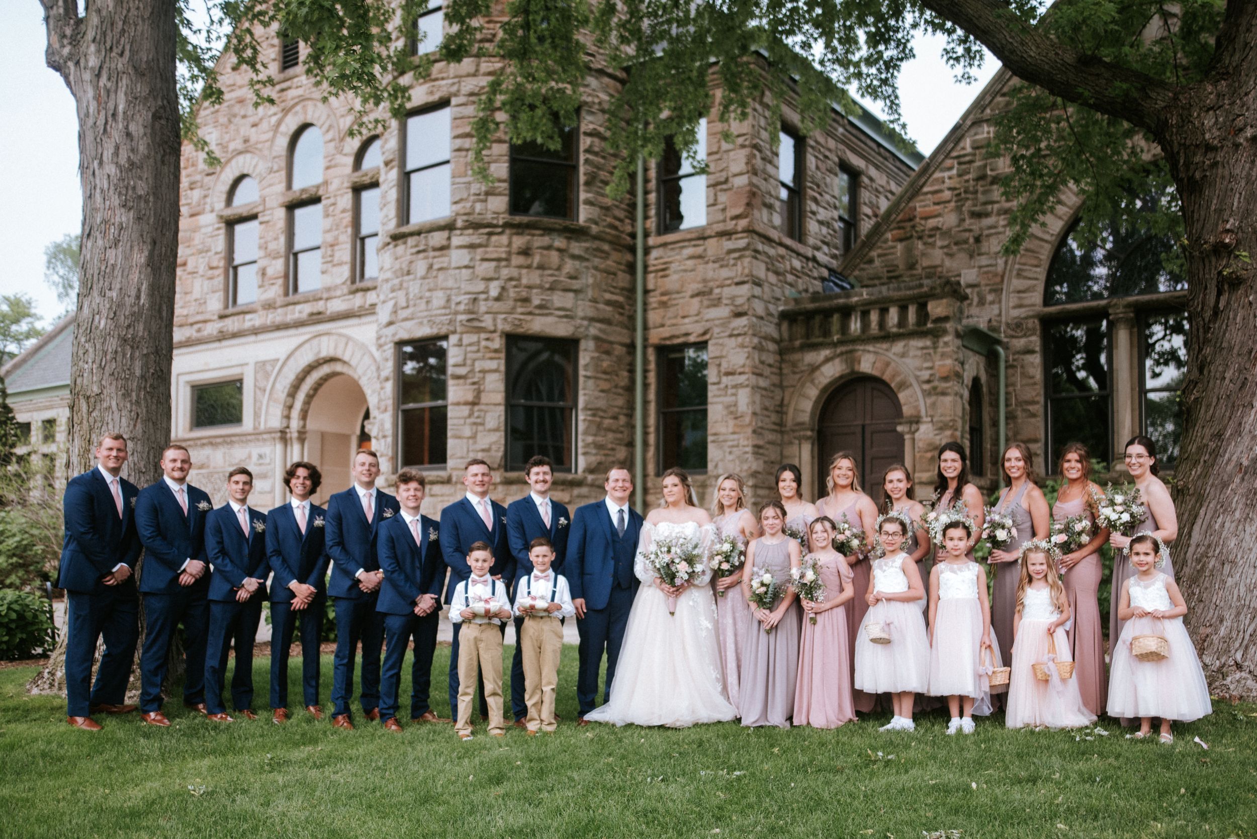 Wedding party in pink dresses and navy suits posed in front of historic stone college building.