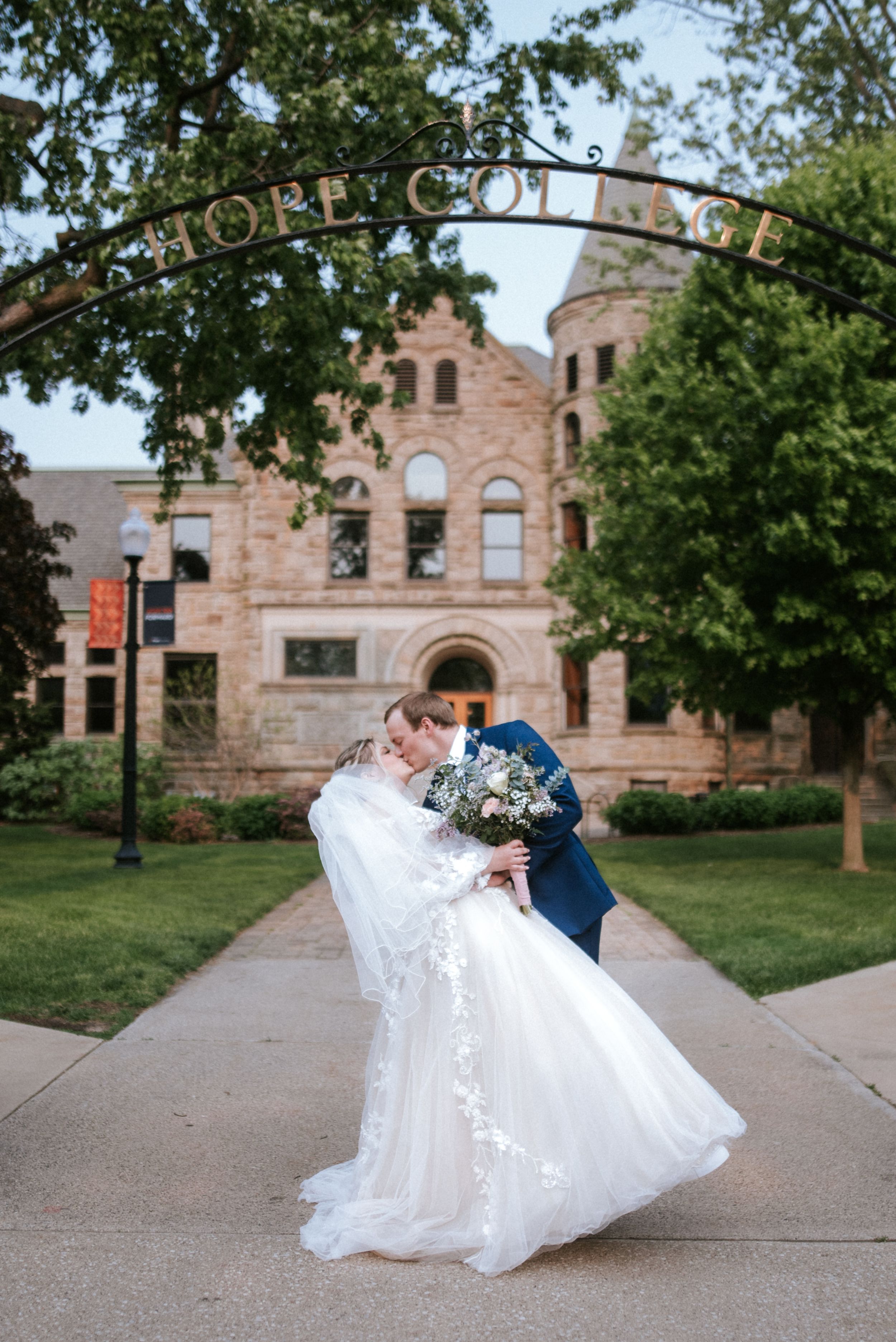 A romantic wedding photo in front of a historic stone college building with ornate architecture and lush trees.