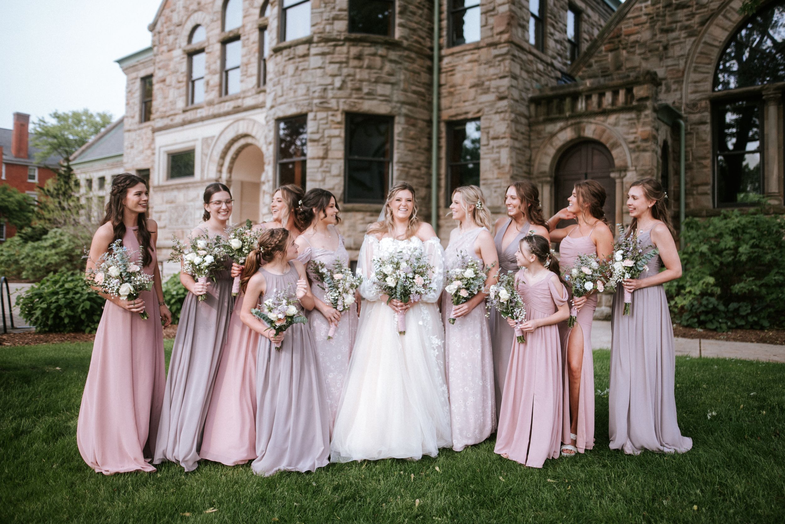 Wedding party in pink bridesmaid dresses poses in front of historic stone castle-like building with bouquets.