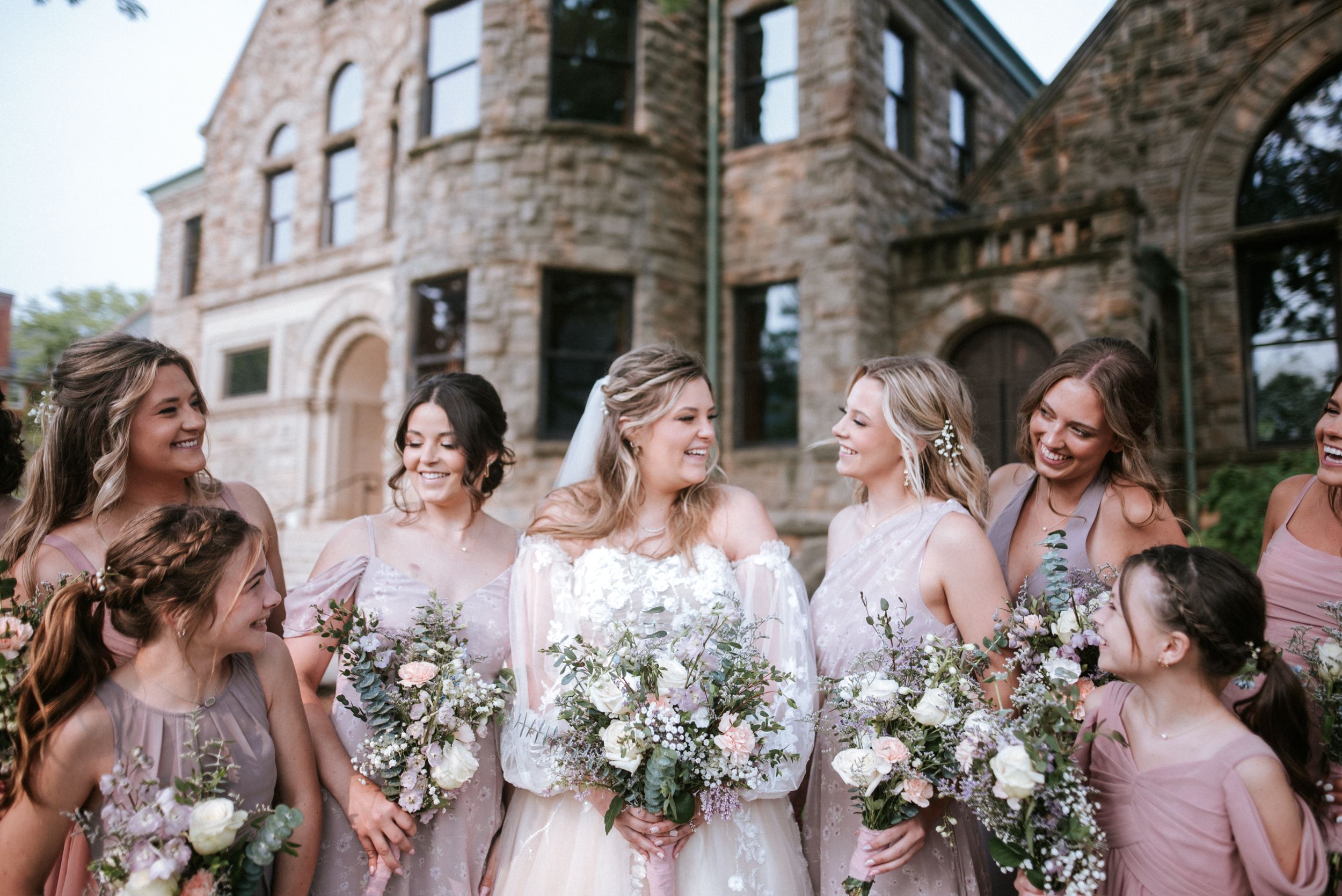 Bridal party shares joyful moment together while holding floral bouquets outside stone building.