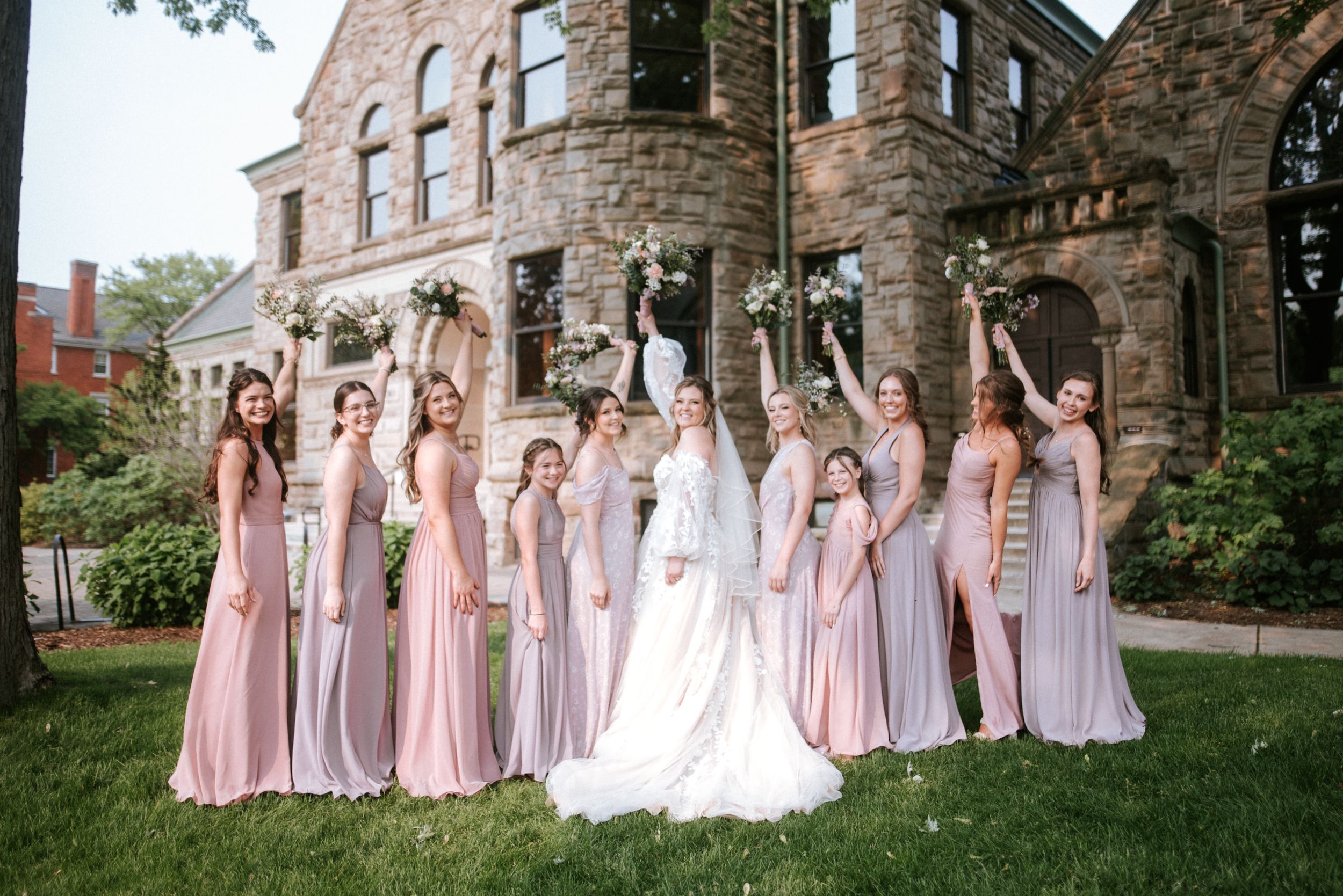 Celebration pose of bridal party in pink dresses on lawn in front of stone building.