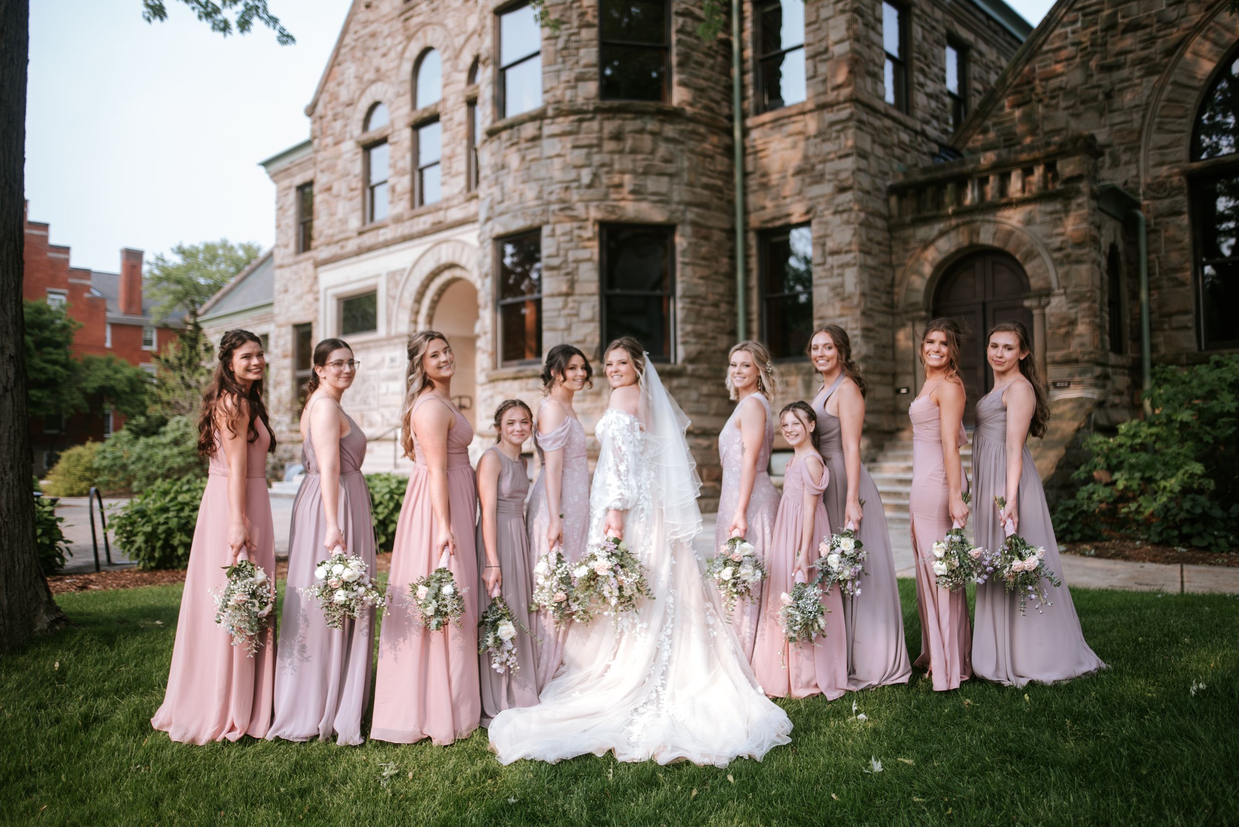 Bridesmaids in pink dresses pose with bride on lawn in front of stone mansion.