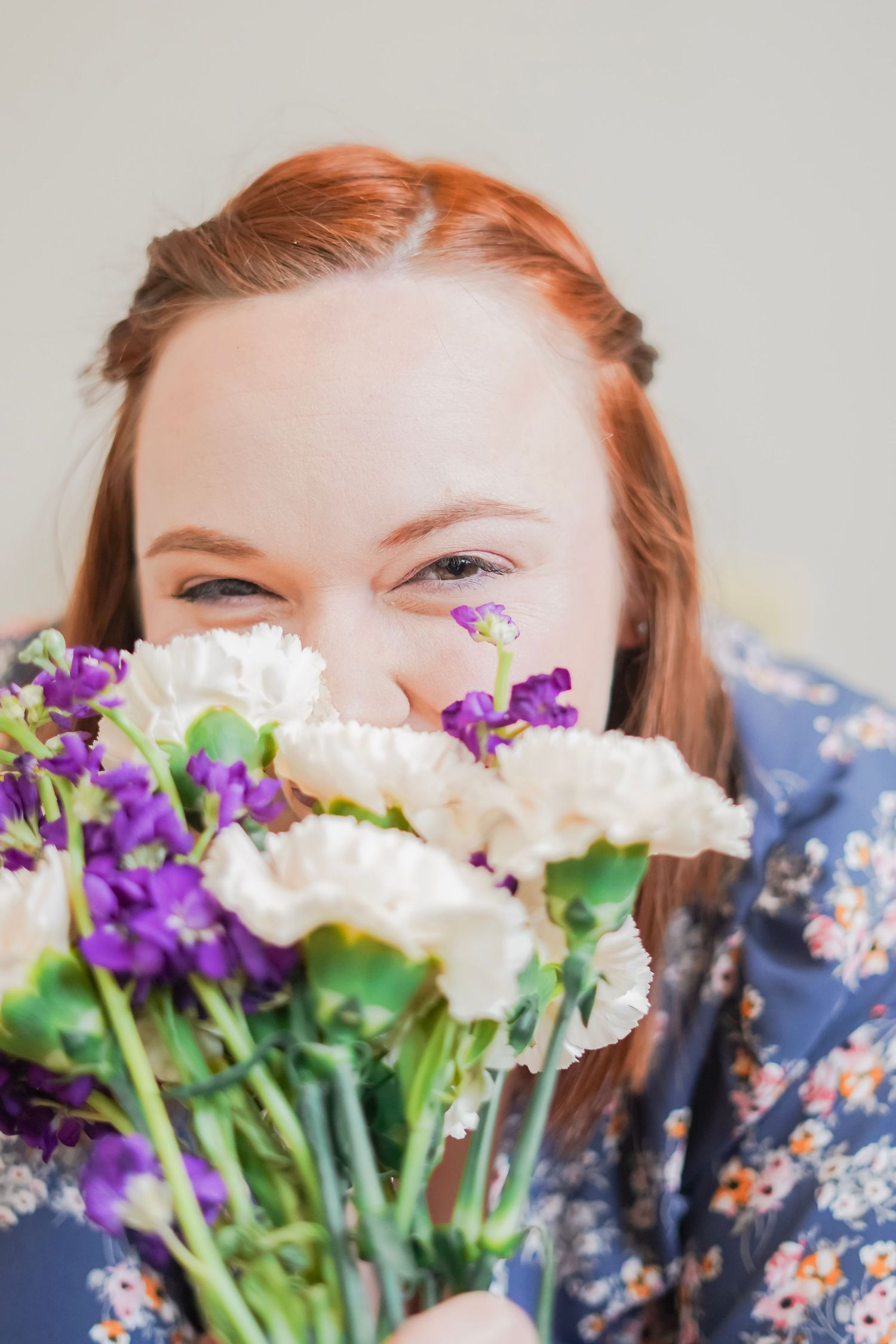 Redhead woman smiling behind a bouquet of white and purple flowers indoors.