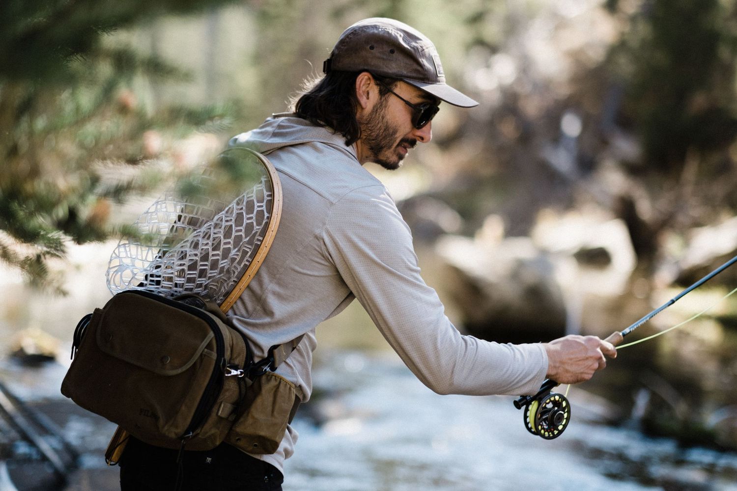 Fly fisherman casting line into mountain stream with scenic forest backdrop.