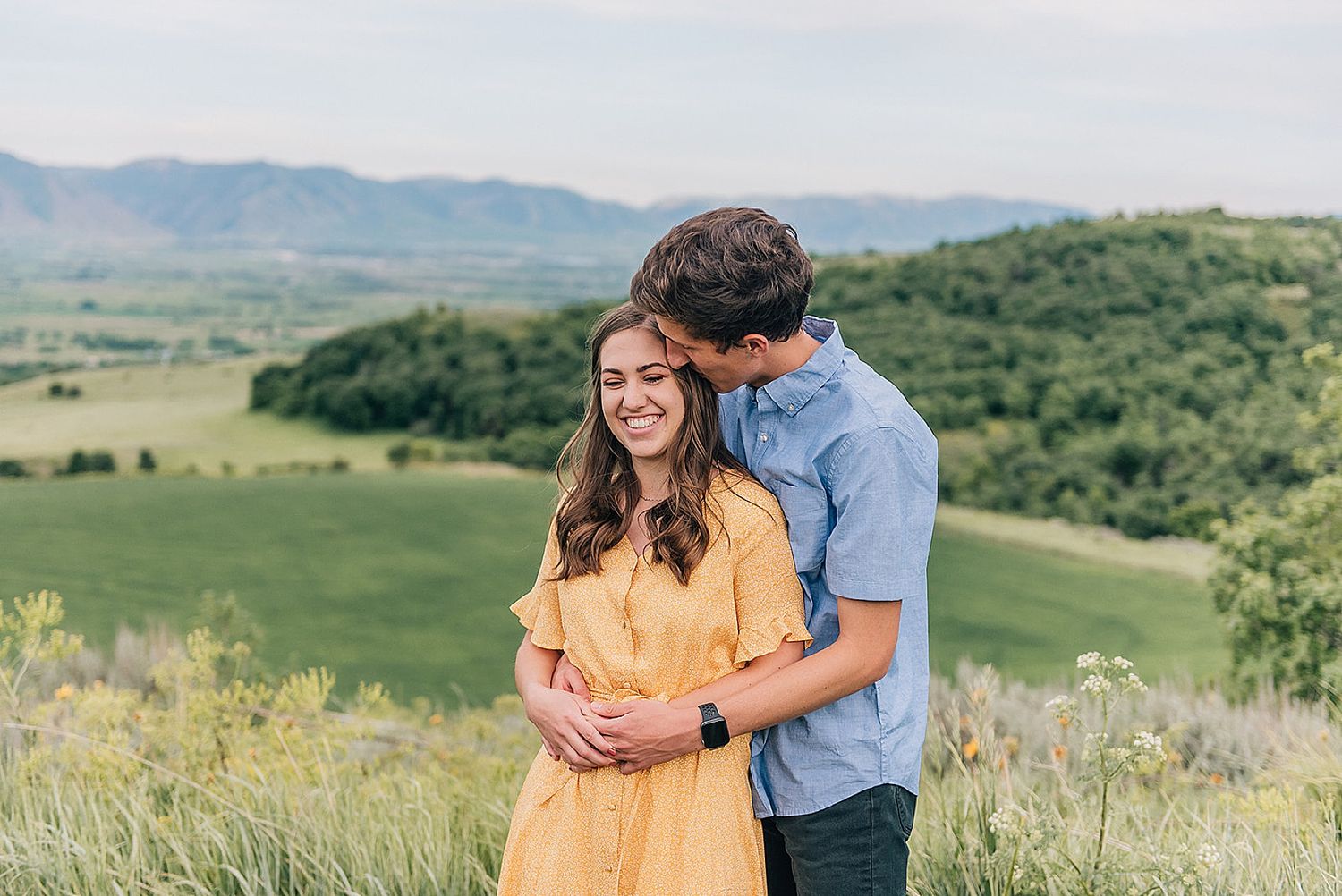 Couple embracing in yellow dress and blue shirt against mountainous backdrop.