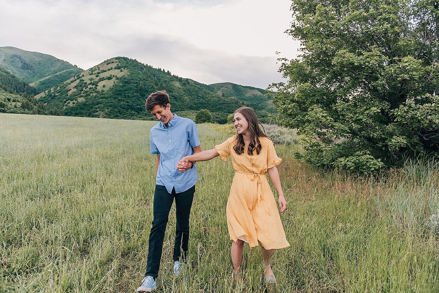 Playful couple walking through tall grass with mountains in distance.