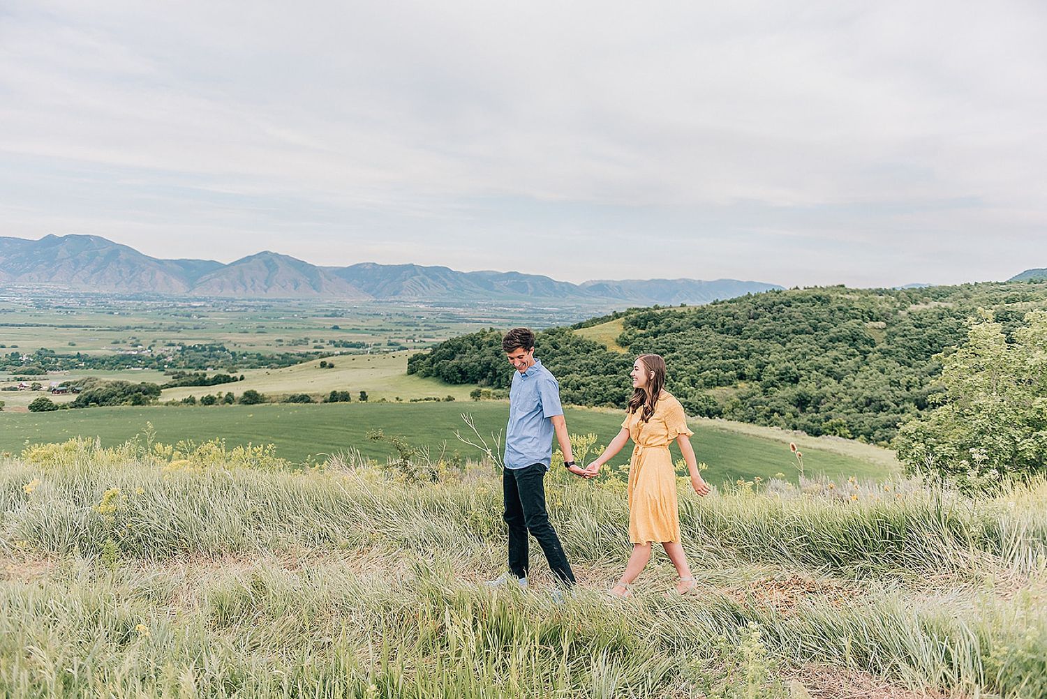 Couple walks together through tall grass with mountains and valley views in background.