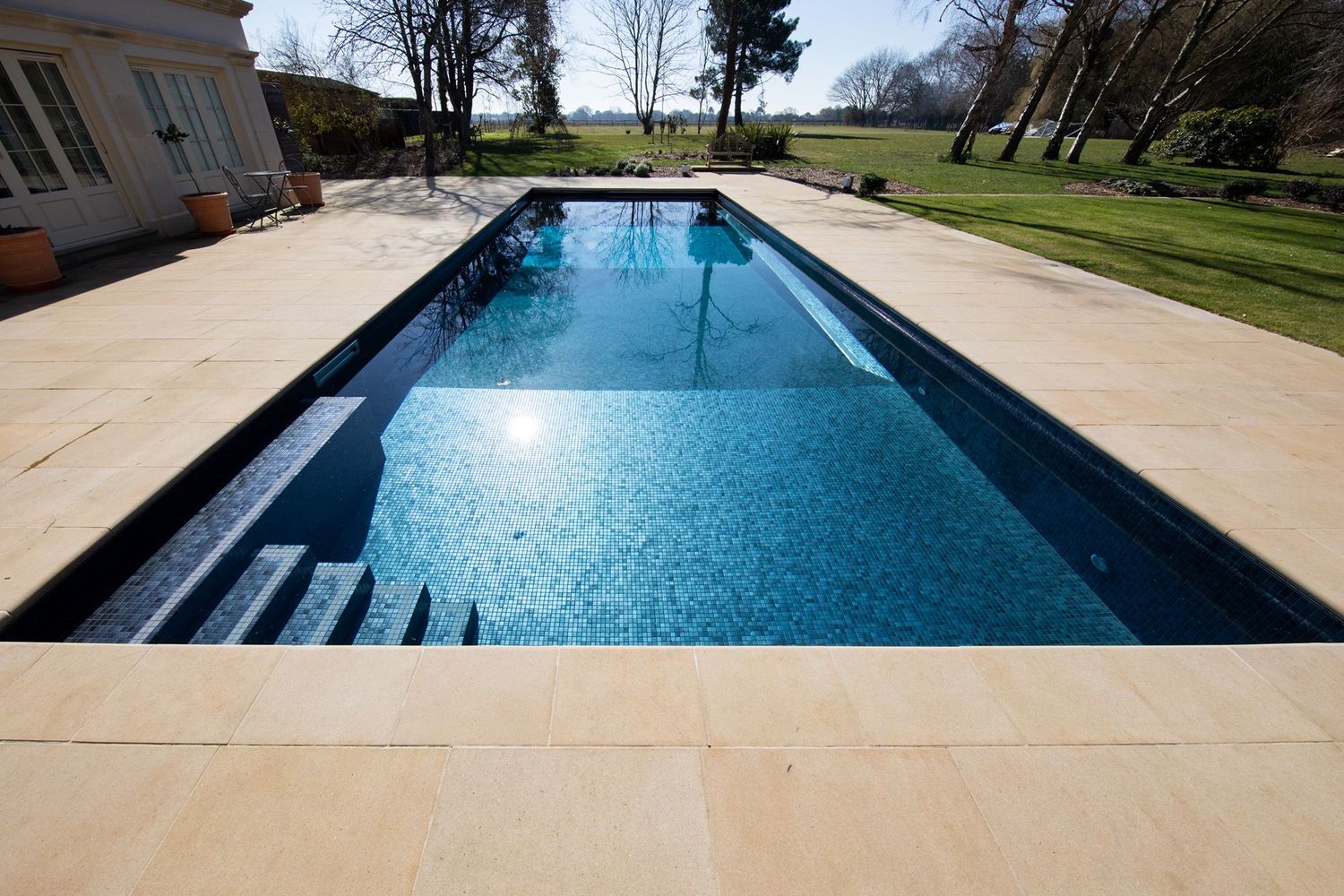 Long rectangular lap pool with dark blue tile and limestone deck surrounded by manicured lawn.