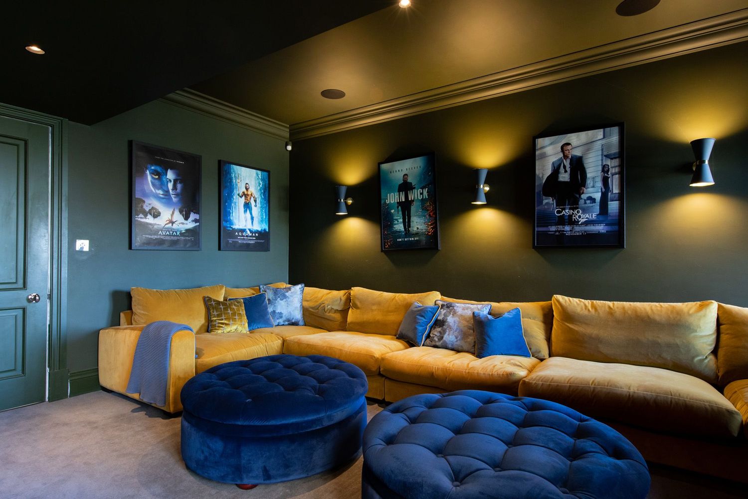 Home theater room with tan sectional sofa, blue ottoman, and movie posters illuminated by wall sconces.