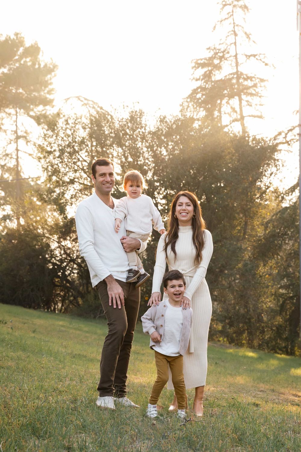 Joyful family of four standing on grassy field at sunset, parents holding children in warm light.