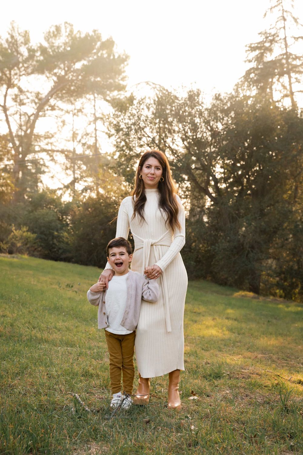 Mother in white dress standing with excited young son in golden hour park setting.
