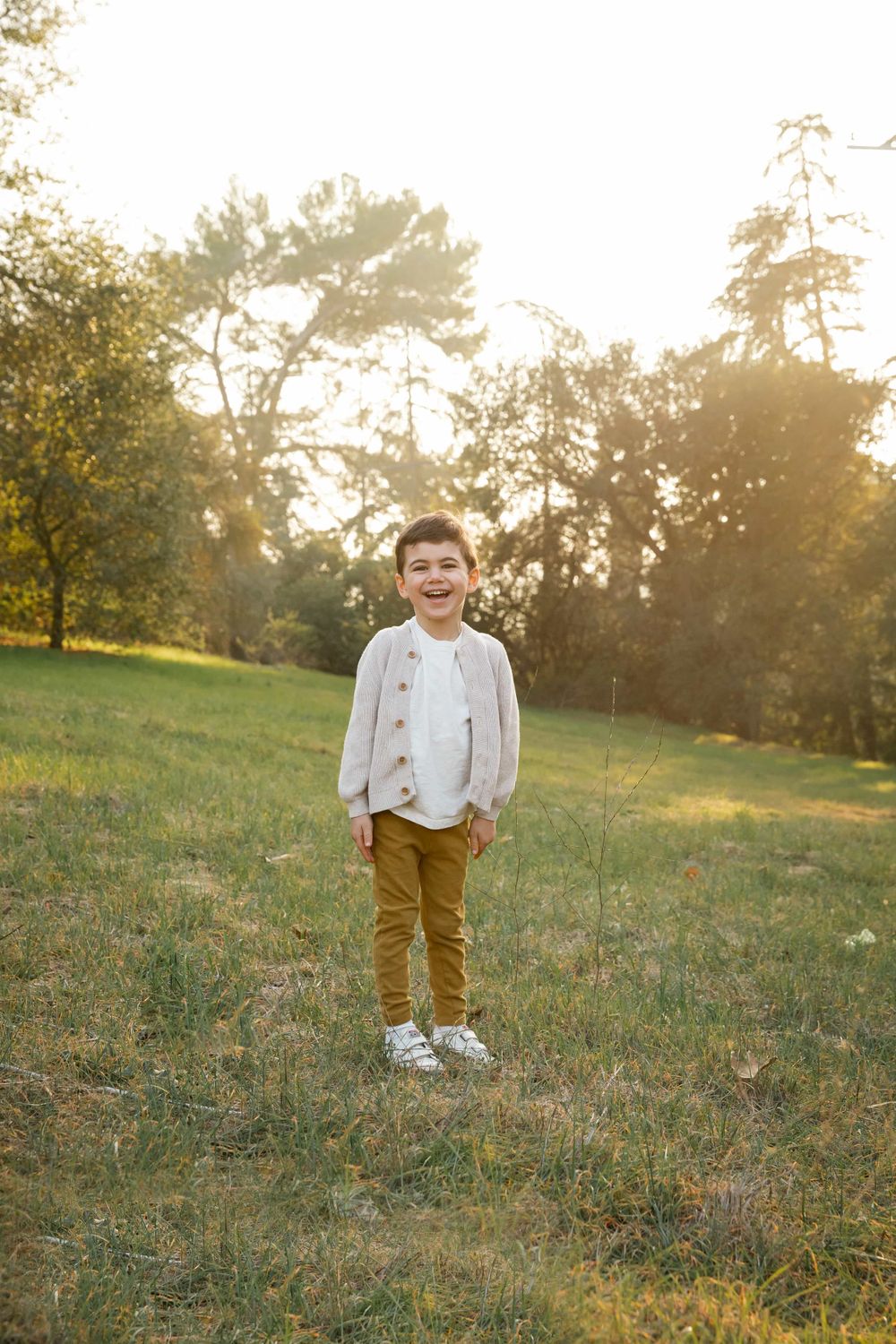 Happy young boy in beige cardigan and mustard pants laughs in a golden sunlit park.