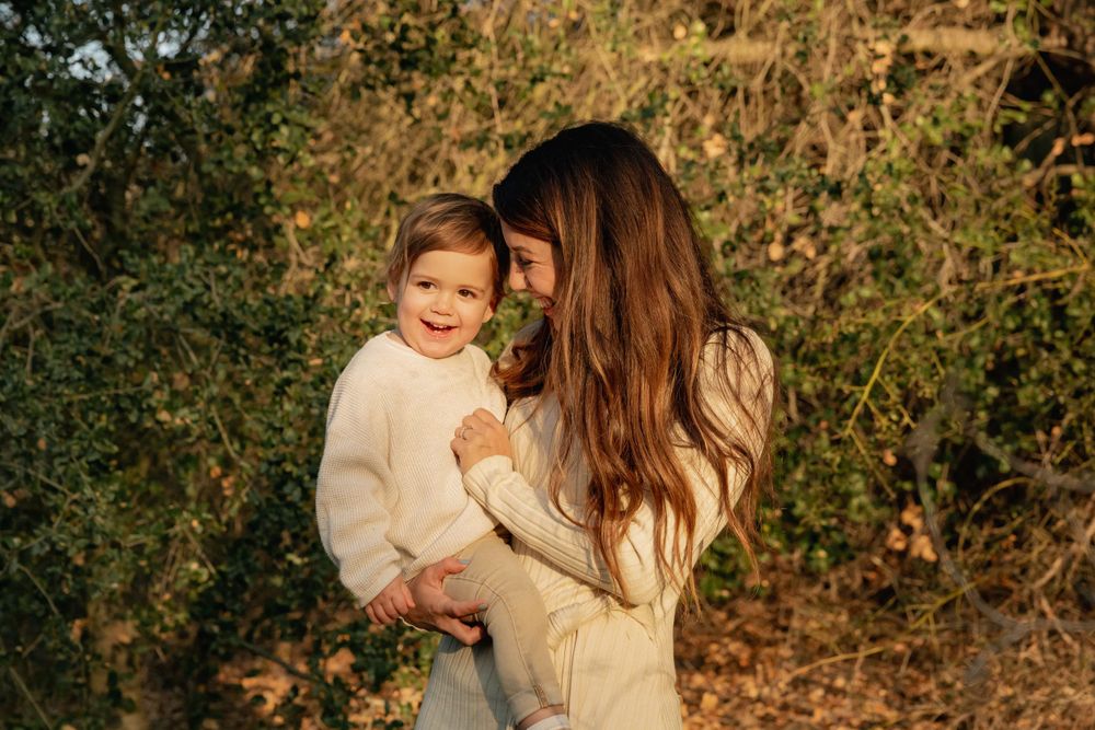Mother with long brown hair holds smiling toddler in white sweater outdoors in warm light.