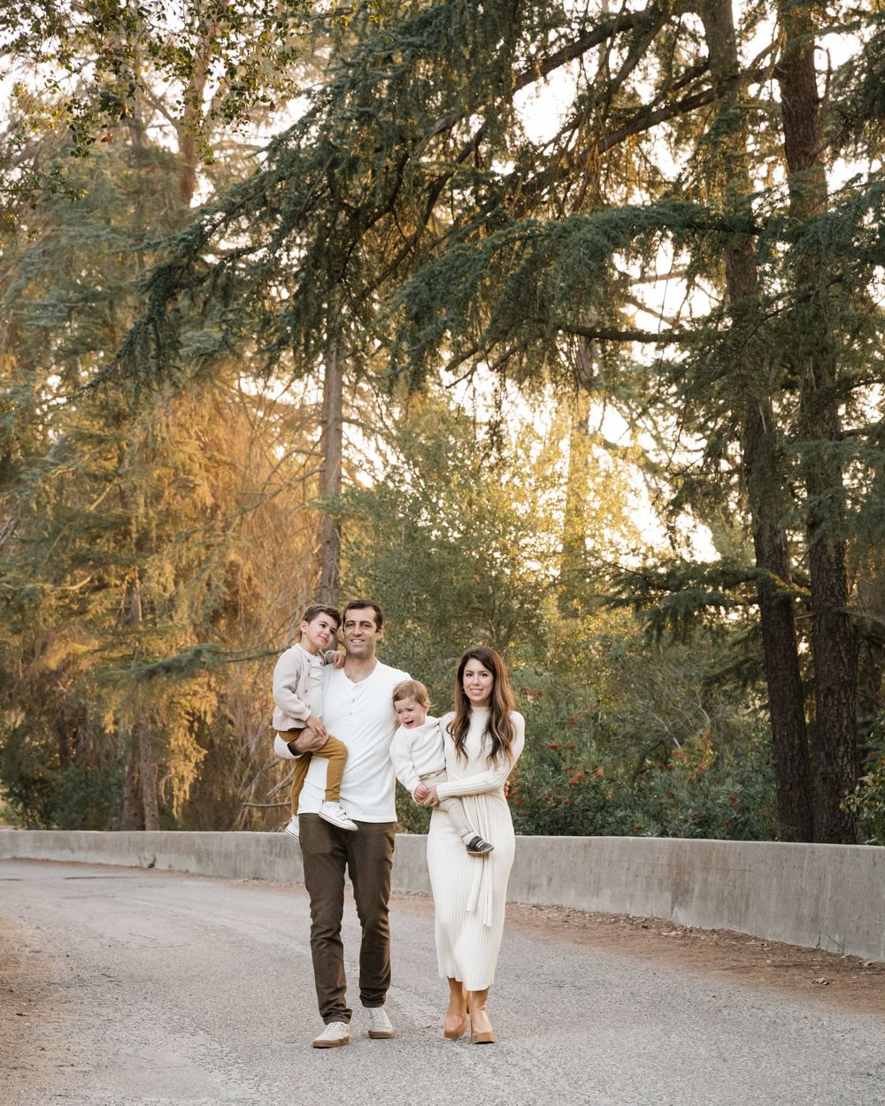Family of four walks on gravel path surrounded by tall trees in golden evening light.