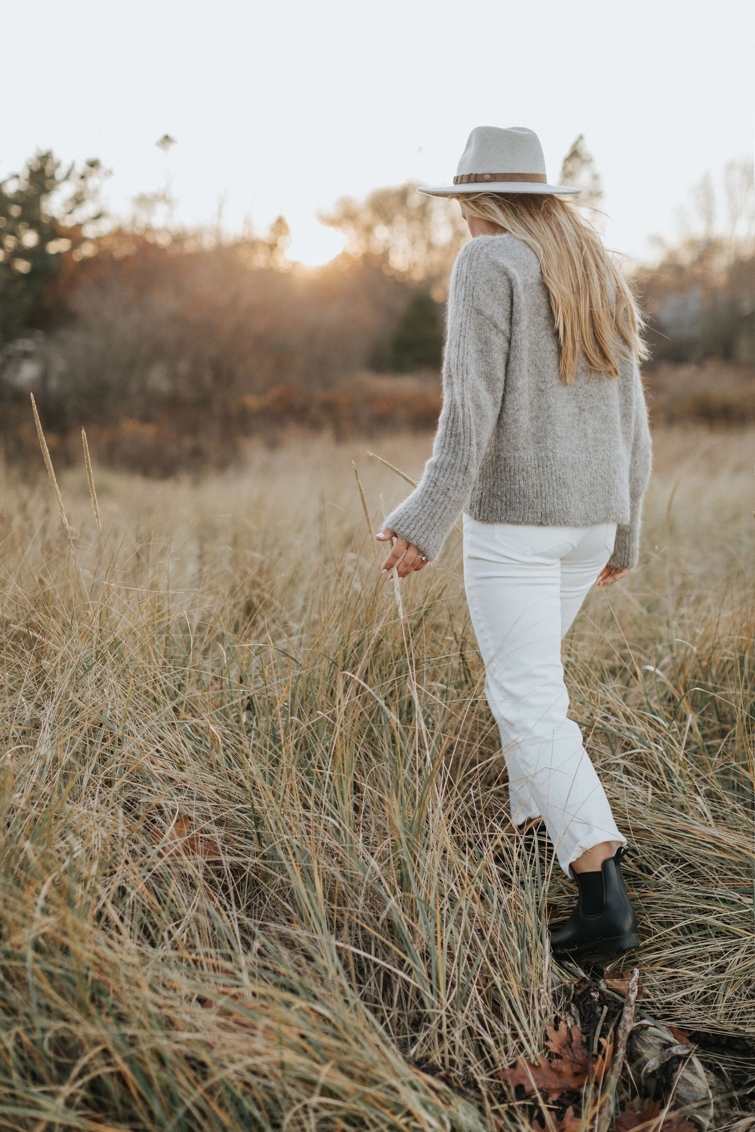 Person wearing grey sweater and white pants walking through tall grass at sunset.