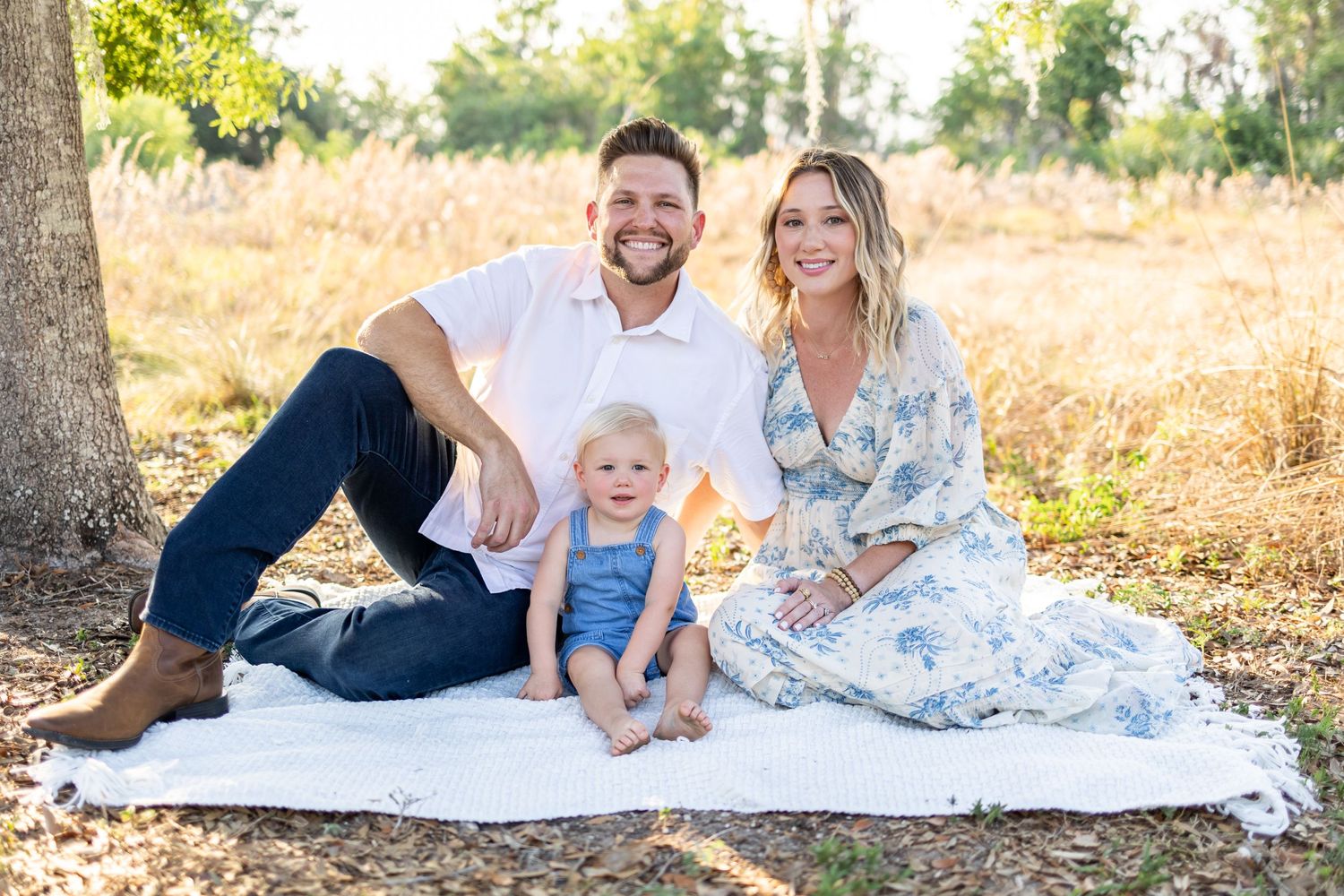 Family relaxes on white blanket under shady trees during outdoor portrait session.
