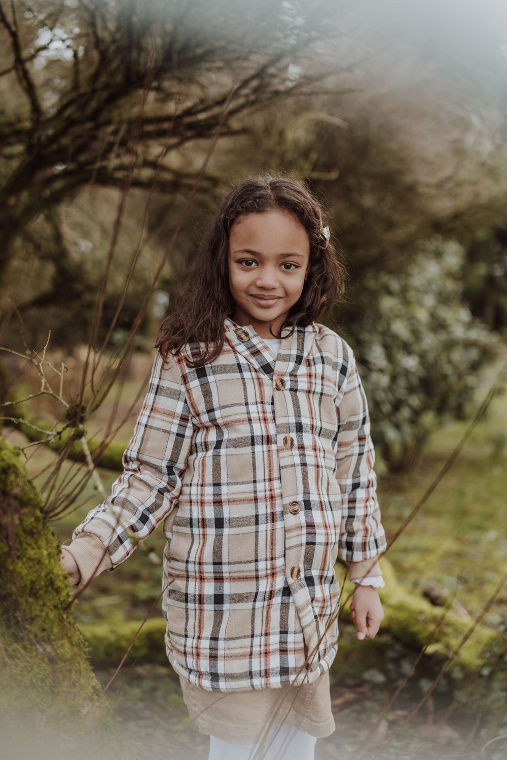 A child in a plaid button-up coat stands outdoors against a misty natural background.