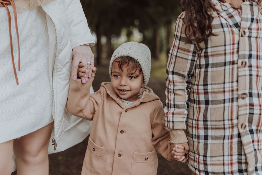 A young child in a beige coat holds hands with family members while walking outdoors.