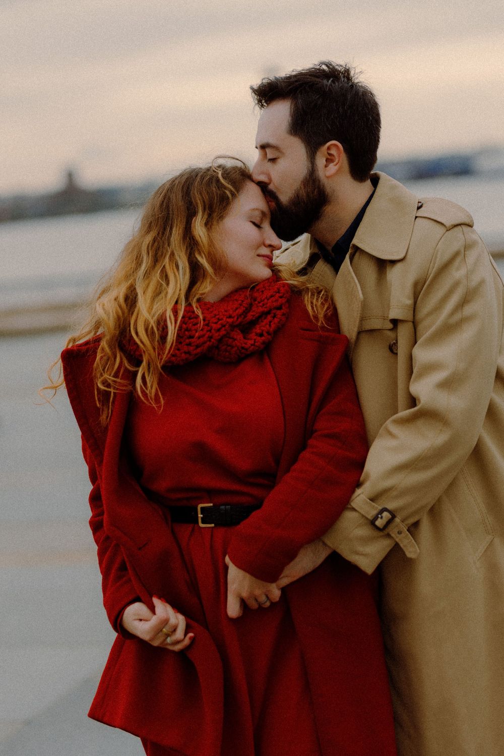 Romantic couple in contrasting coats share tender moment against cloudy backdrop.