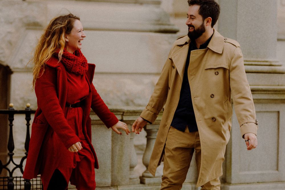 Couple in coordinated outfits walking playfully along urban building exterior.