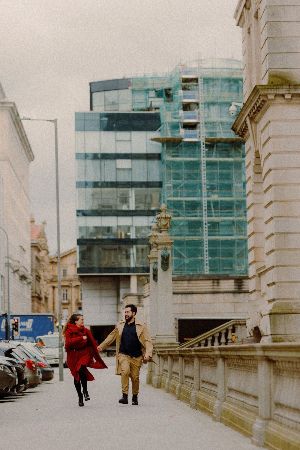 Couple in red and beige coats walking near modern glass building with scaffolding.