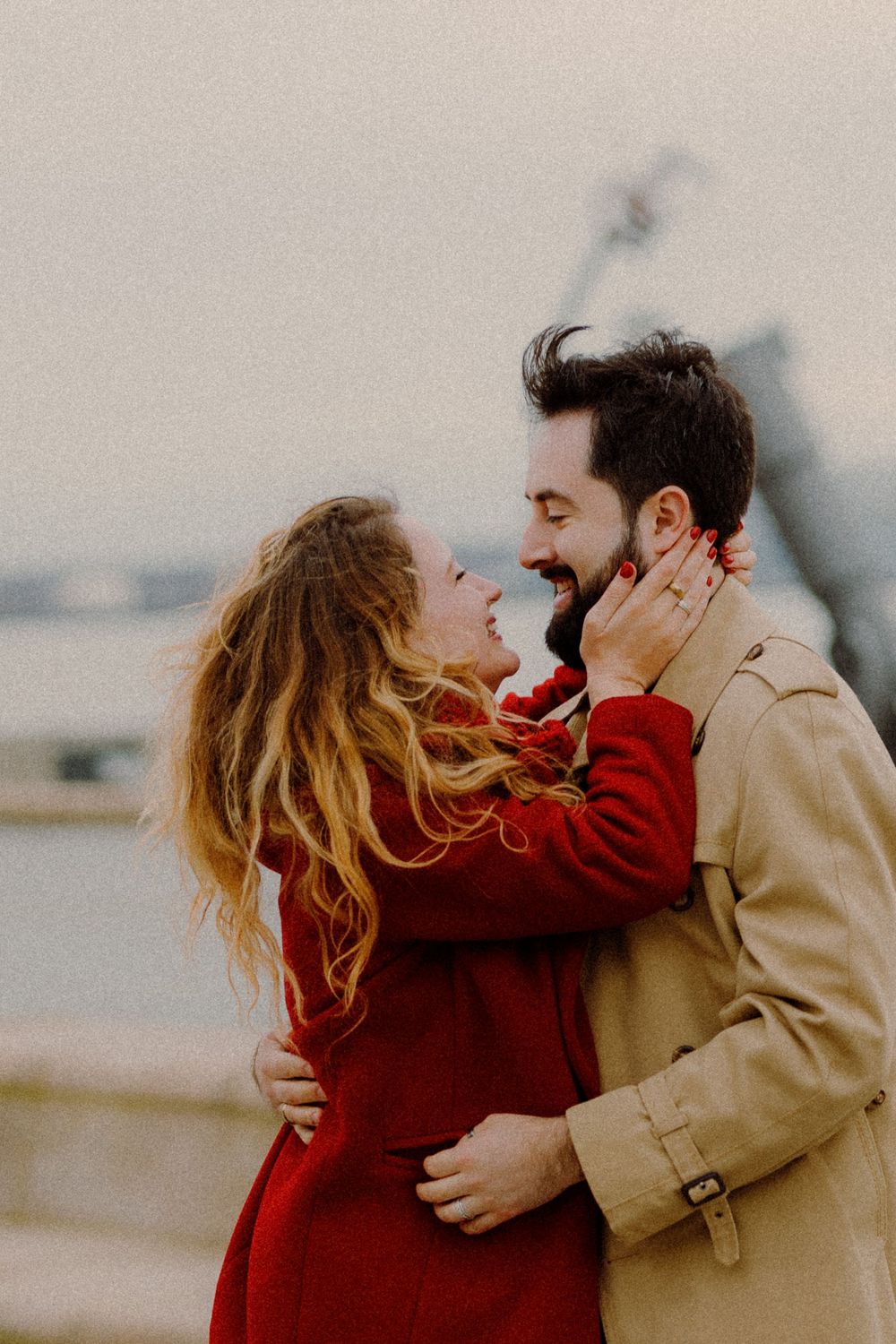 Couple sharing intimate moment outdoors, with woman in red and man in beige coats.