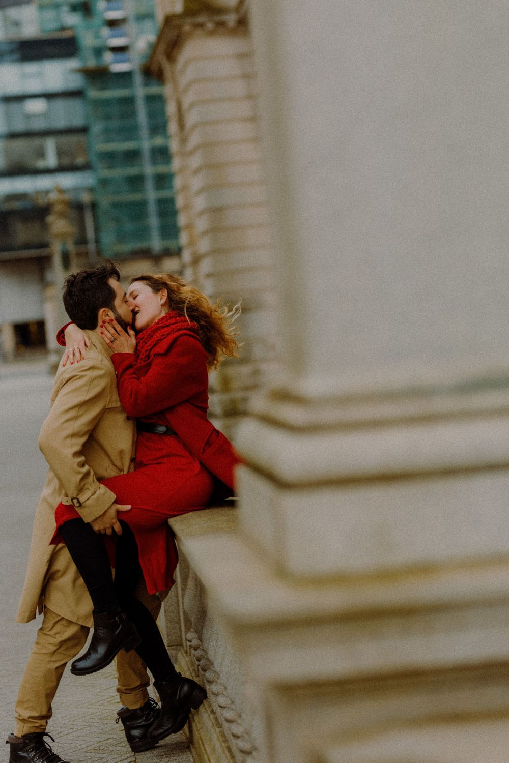 A romantic couple in tan and red coats share a passionate embrace against an urban backdrop.