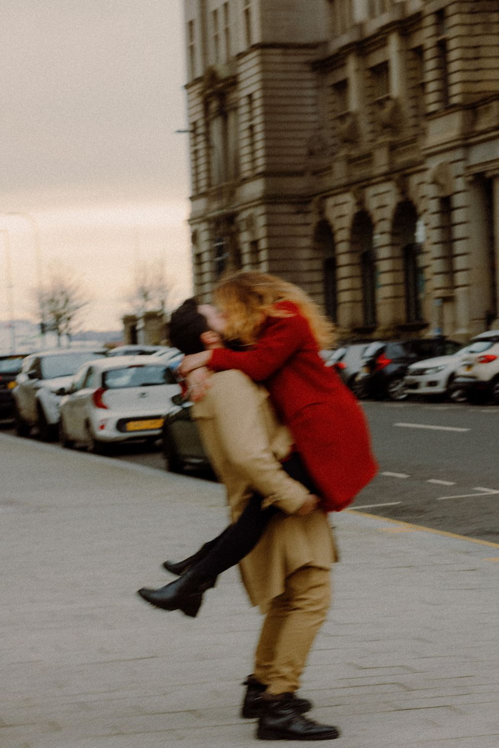 A couple in contrasting beige and red coats share a playful lift moment on a city street.