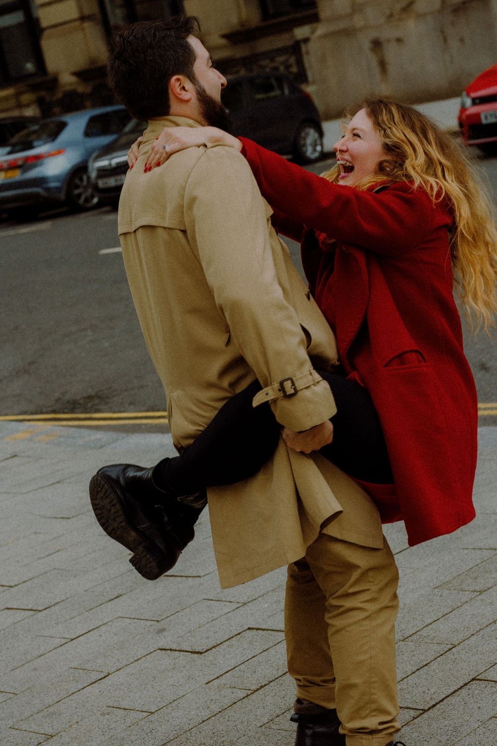 A joyful couple in tan and red coats dancing and laughing together on a city sidewalk.