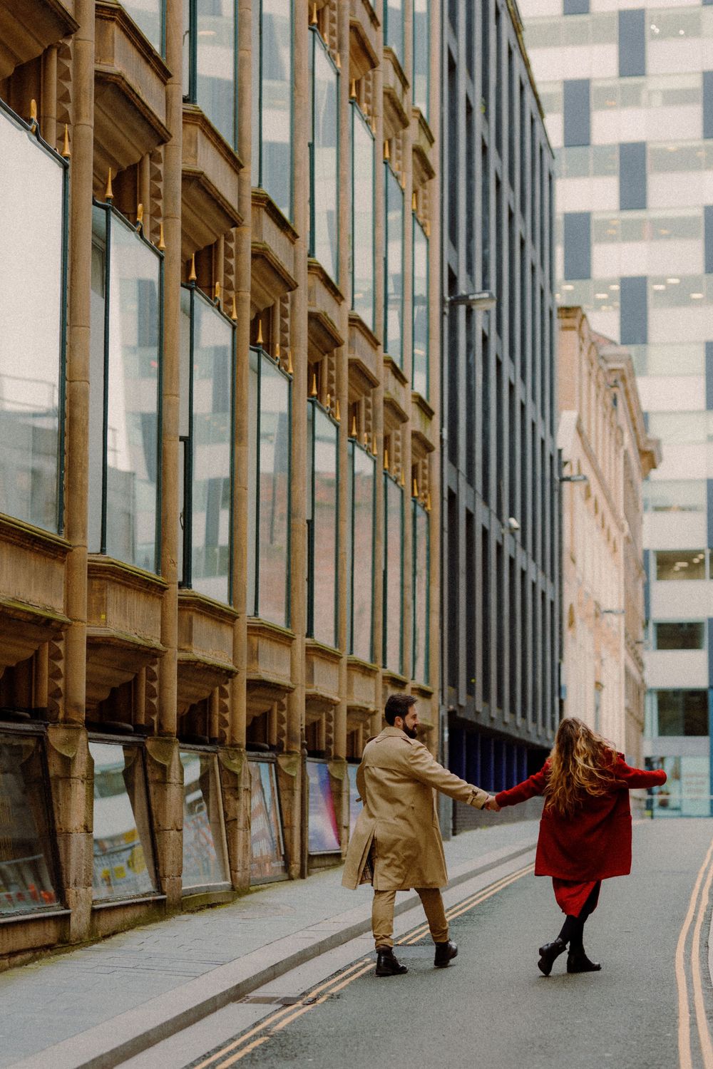 Two people in red and tan coats hold hands while walking past modern glass buildings.