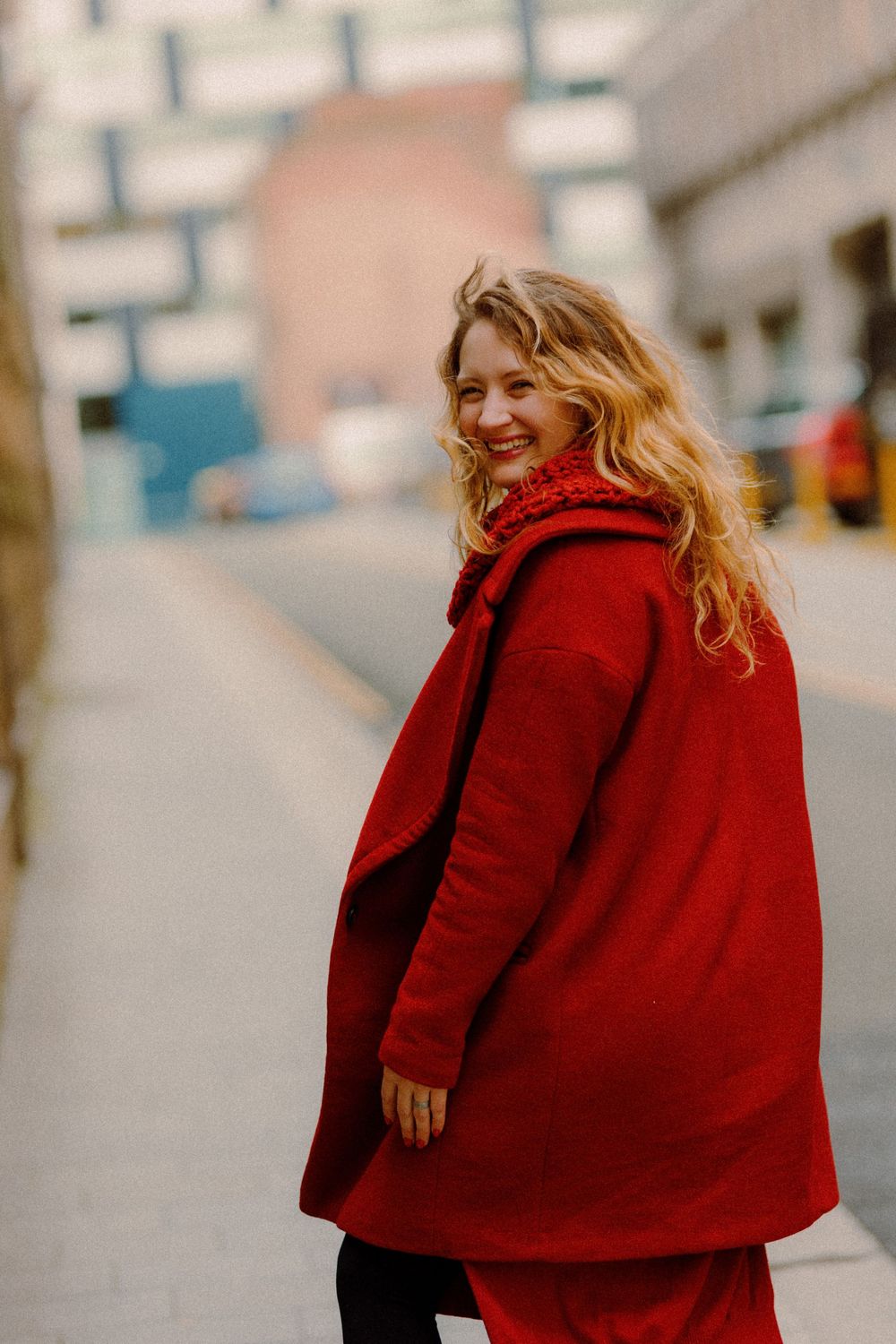 A person in a vibrant red coat smiles while standing on an urban street.
