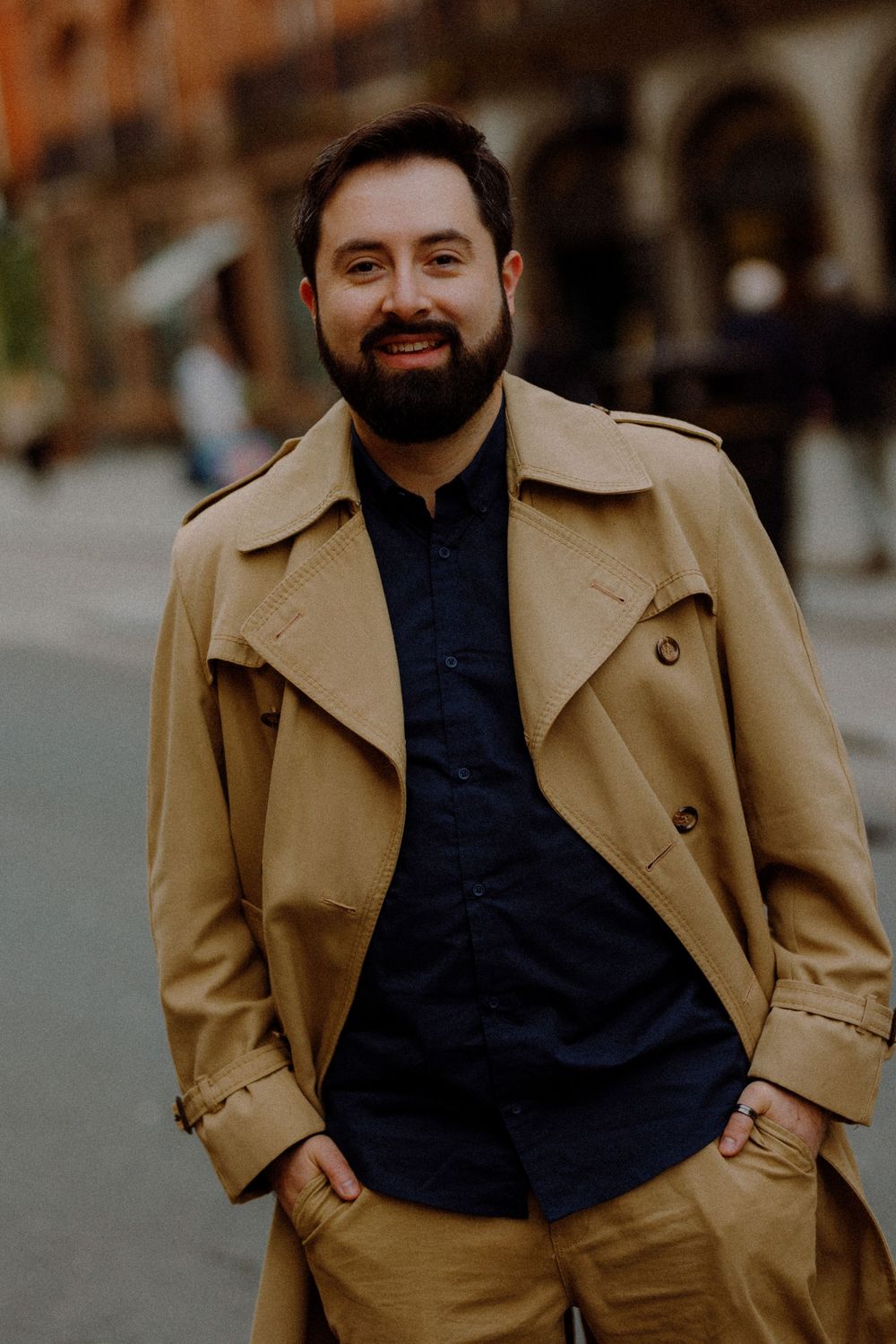 A person in a tan trench coat and navy shirt smiles at the camera on a city street.