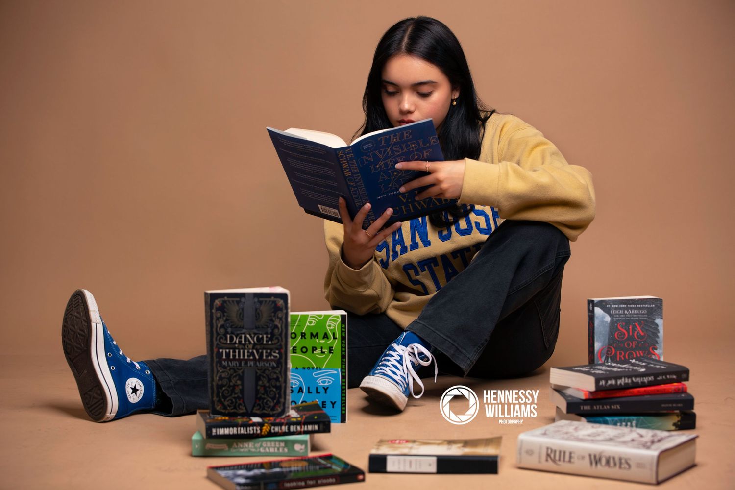 Person in yellow sweater reading surrounded by stacked books on a brown background.