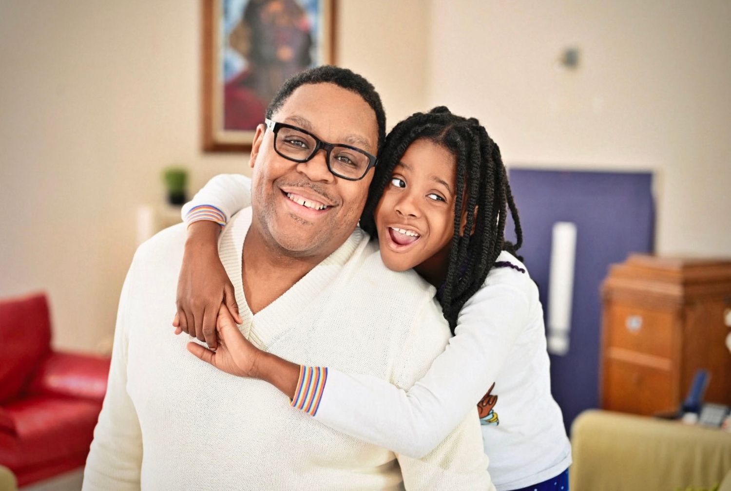 Smiling Black man with glasses shares a warm hug with a young girl in a bright indoor setting.